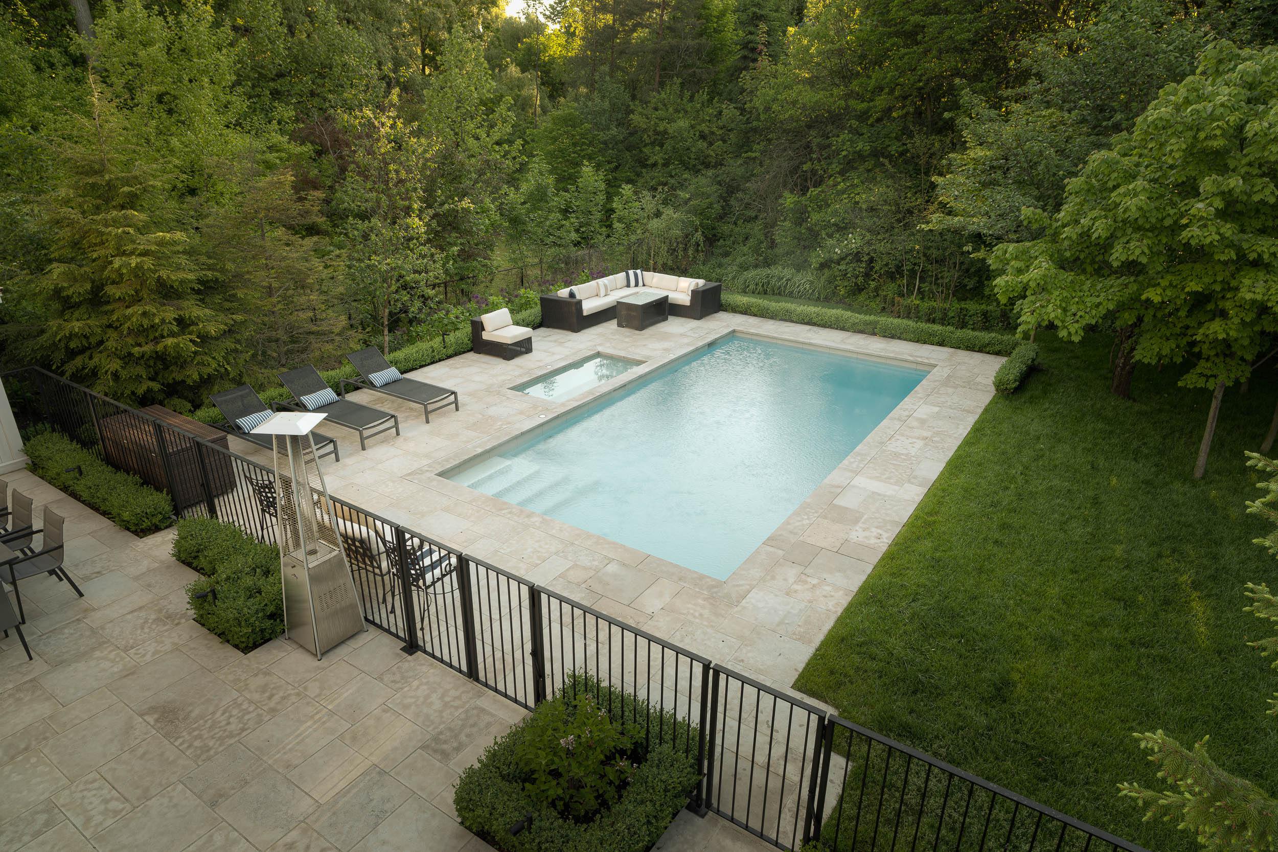 A serene backyard features a rectangular swimming pool, surrounded by lounge chairs, patio furniture, green lawns, and lush trees. Quiet, inviting atmosphere.