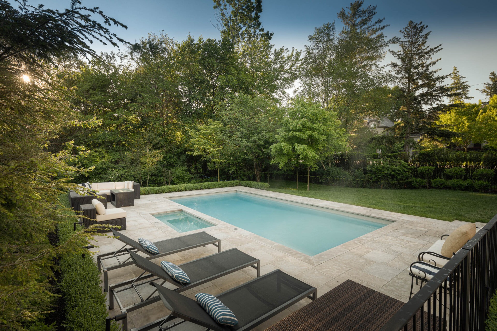 A serene backyard with a rectangular pool, surrounded by lush trees, outdoor seating, and deck chairs under a clear blue sky.