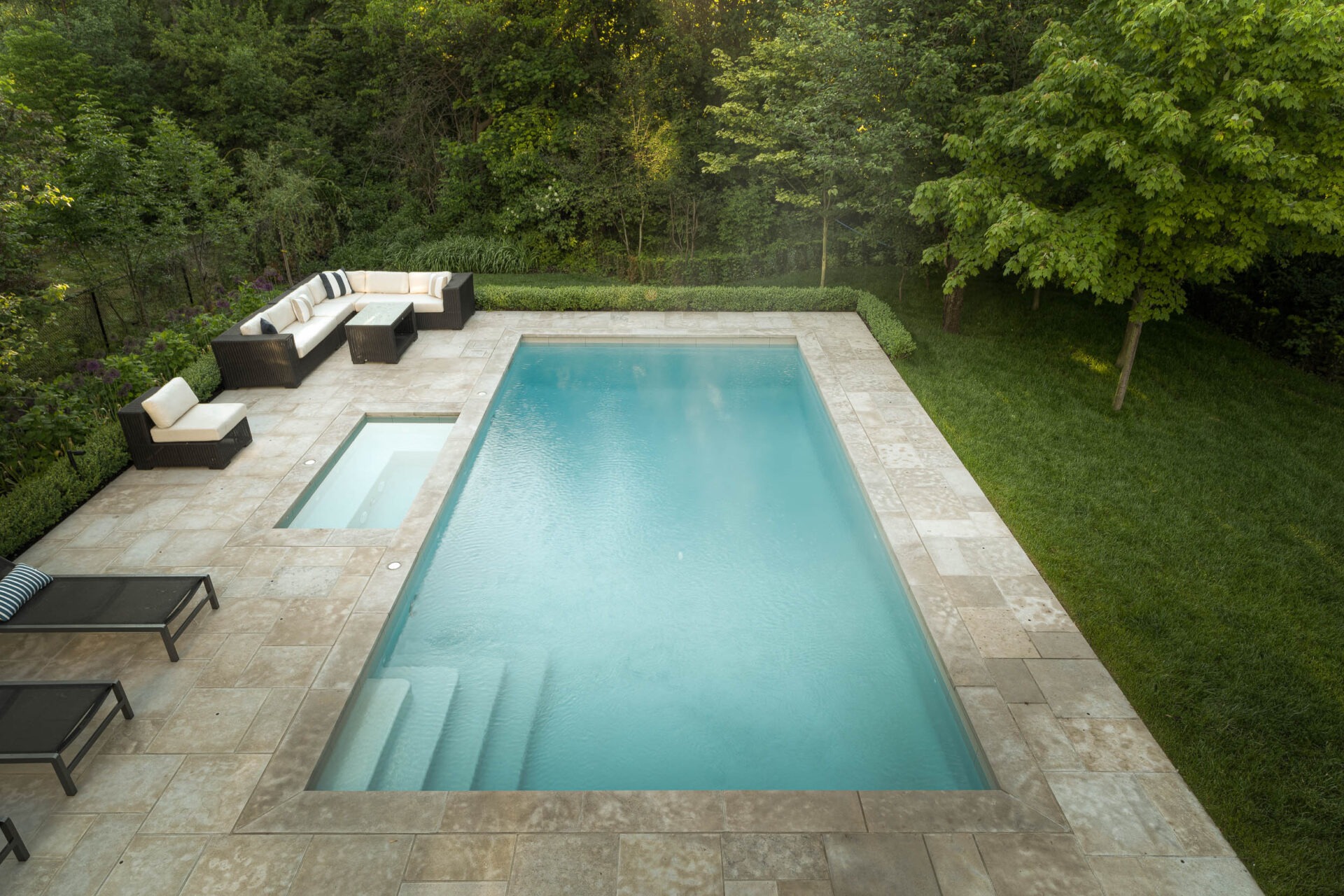 A rectangular swimming pool with adjacent spa, surrounded by trees, patio furniture, and greenery, in a serene backyard setting.