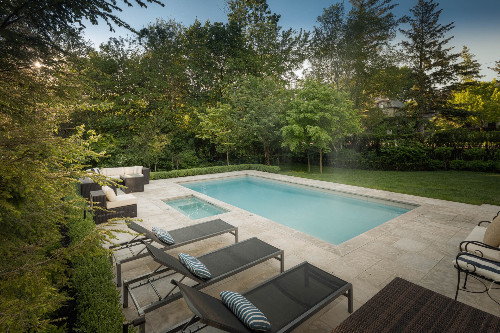 A serene backyard pool area with lounge chairs and cushioned seating, surrounded by lush greenery and trees under a clear sky.