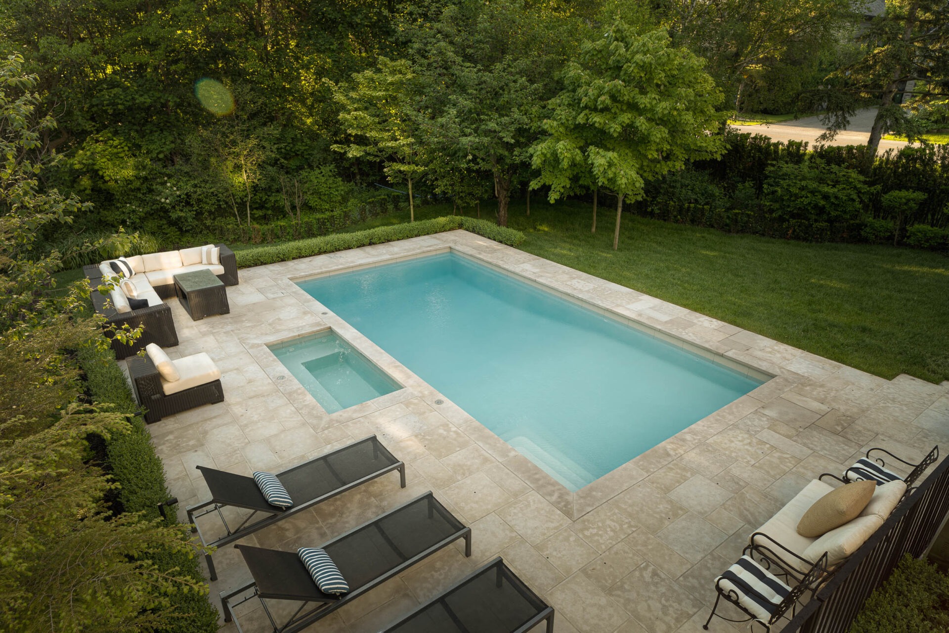 A serene backyard features a rectangular pool with adjacent hot tub, surrounded by comfortable patio furniture and lush greenery.