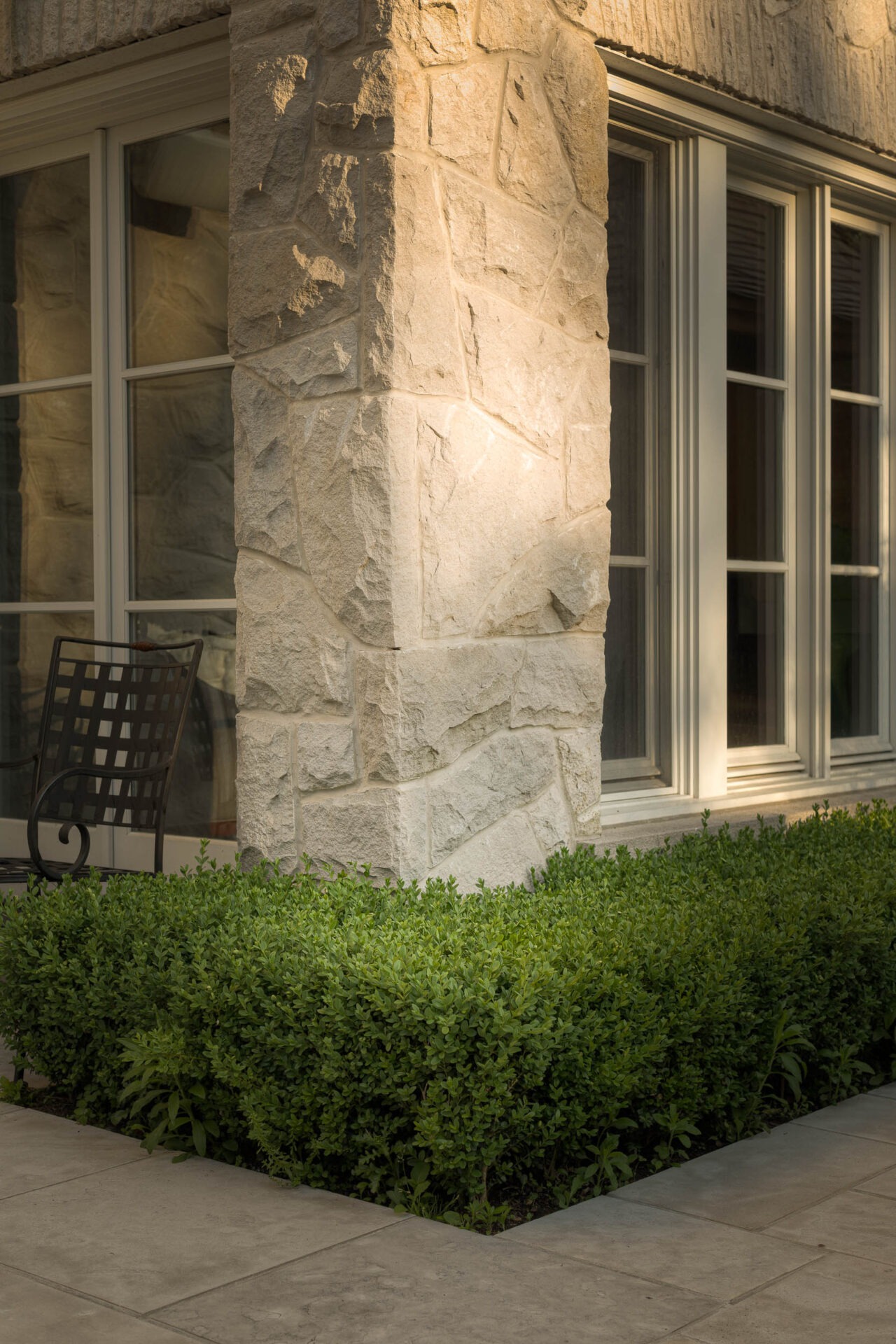 Stone building corner with large windows, a black metal chair, and neatly trimmed green bushes in a serene, sunlit setting.