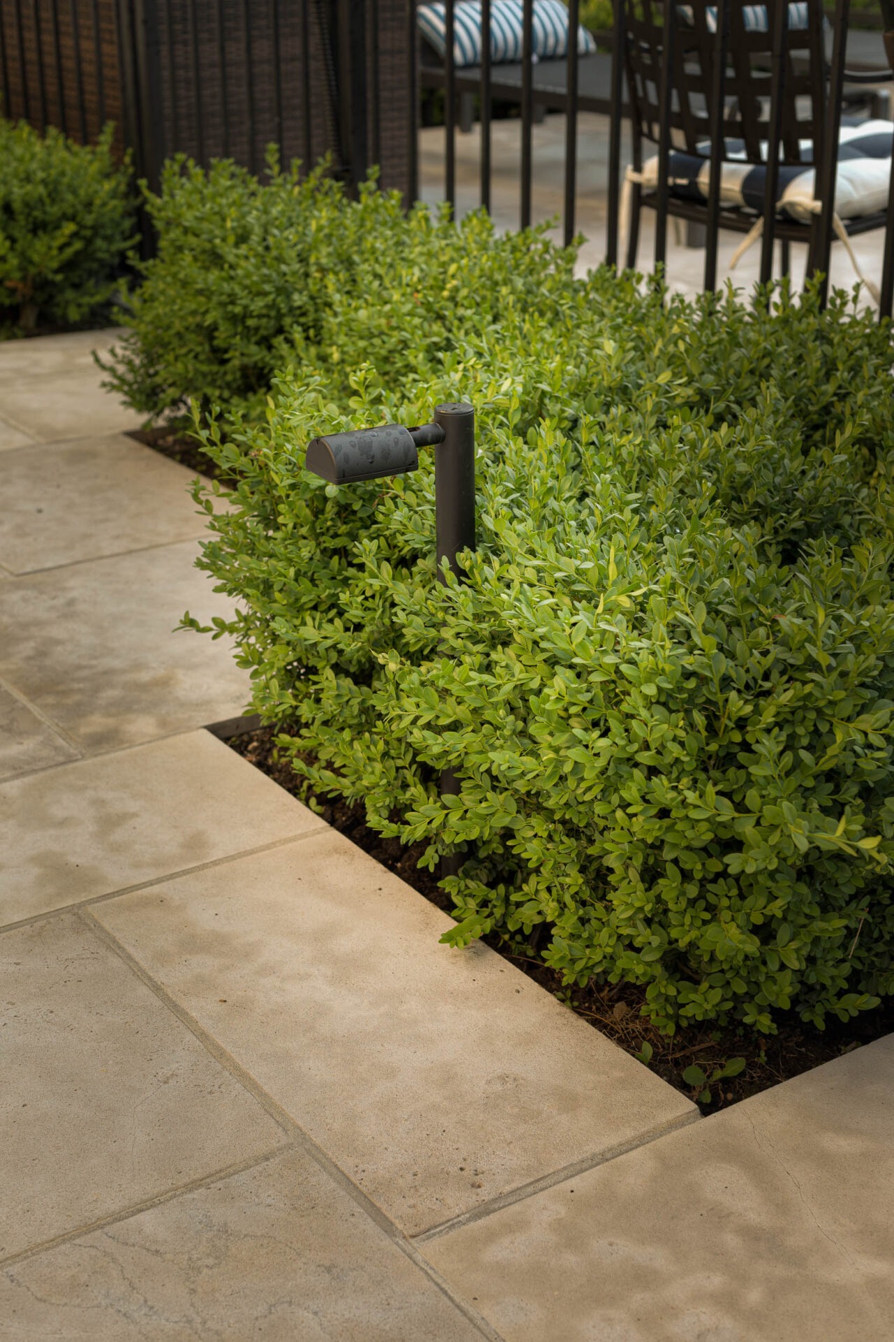 A garden setting with neatly trimmed bushes, stone tile pathway, outdoor seating, and small black garden light, surrounded by a metal railing.