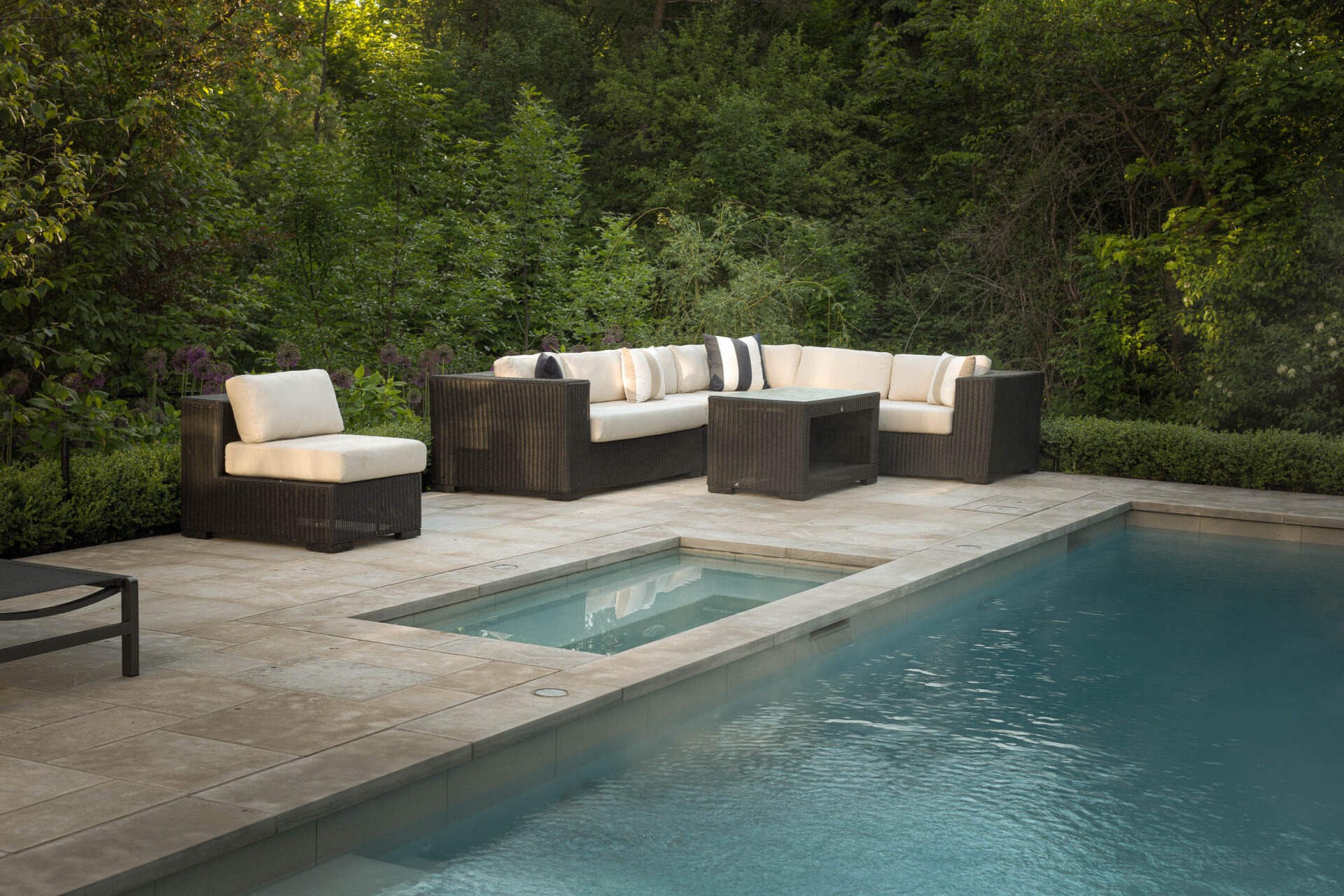 A serene outdoor patio with modern wicker furniture beside a clear blue swimming pool, surrounded by lush green foliage and trees.