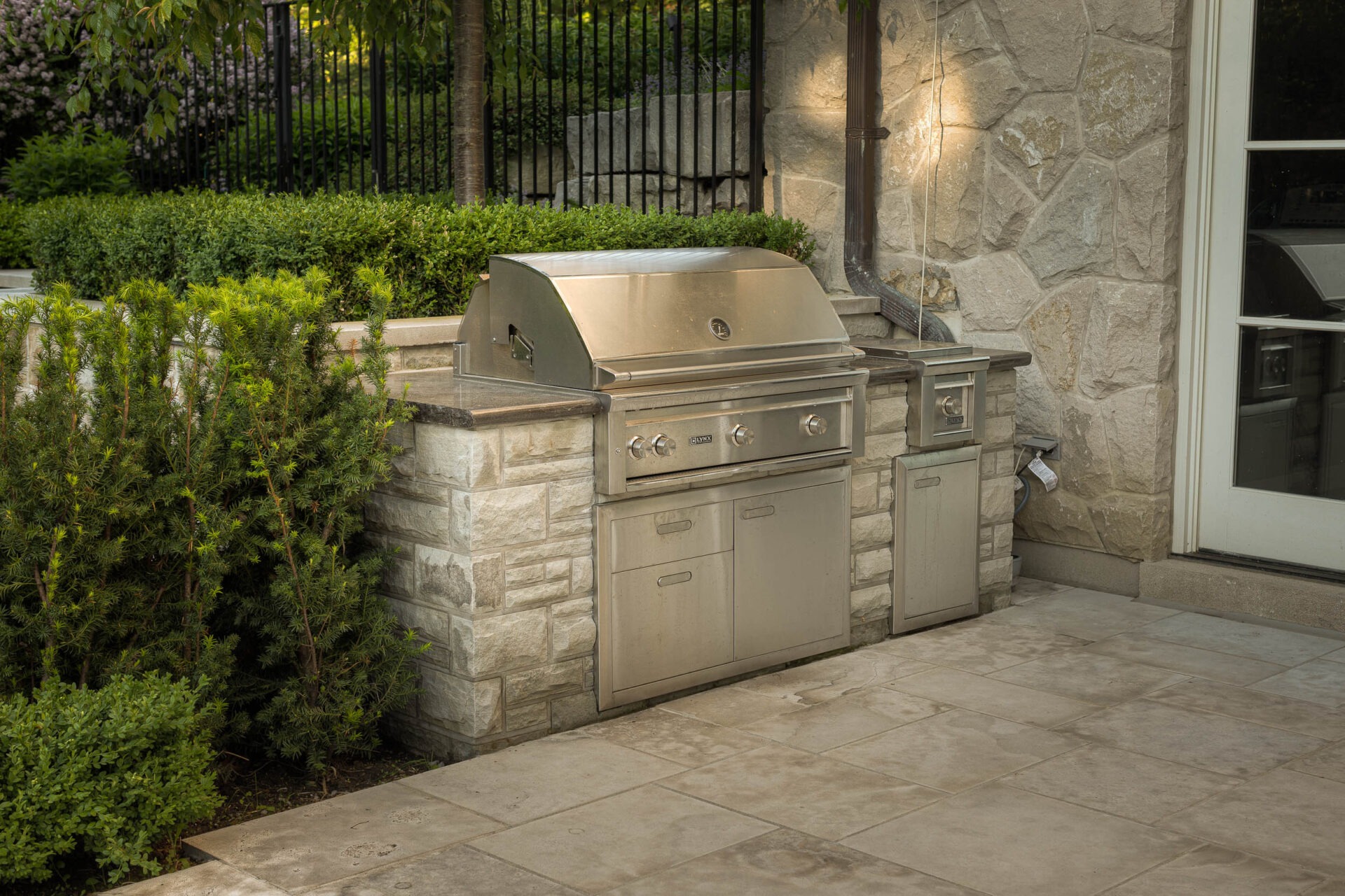 Outdoor kitchen with stone countertops, stainless steel grill, surrounded by lush greenery and stone wall. Paved patio area in front.