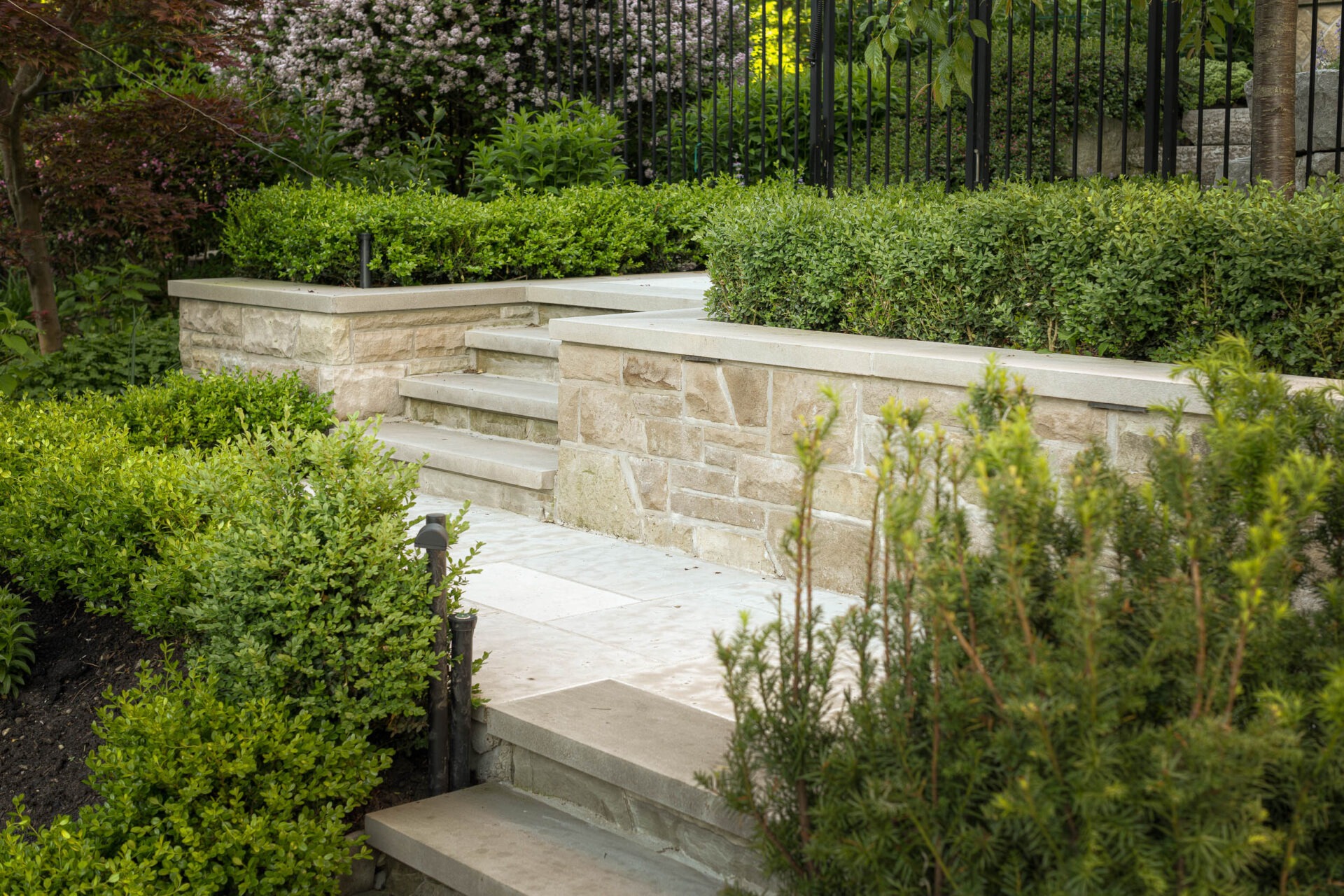 Stone steps with lush greenery and flowering bushes are surrounded by a metal fence in a well-maintained garden setting.