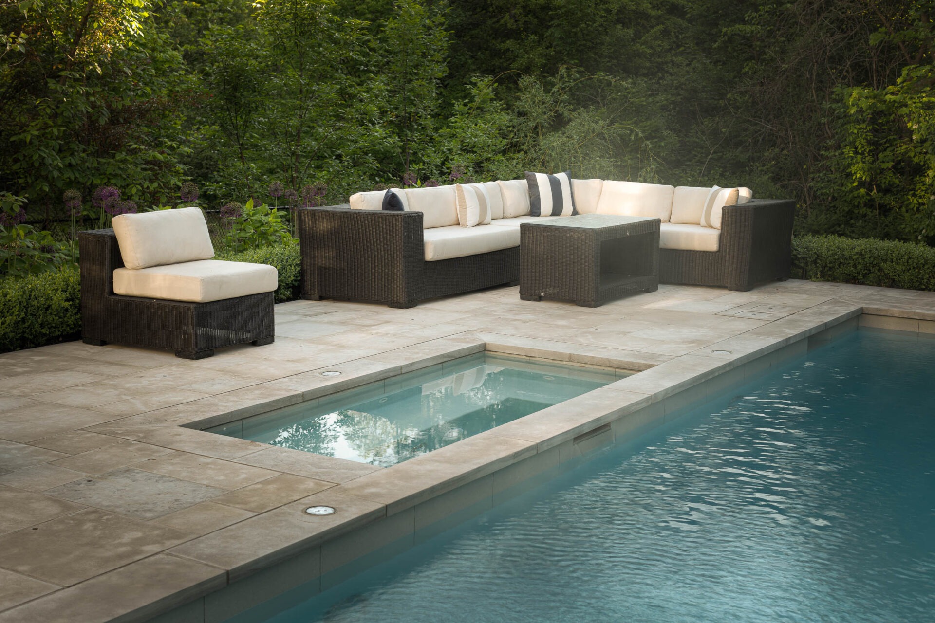 Outdoor seating area with rattan furniture beside a rectangular pool, surrounded by lush greenery and neat hedges. Calm, serene setting.