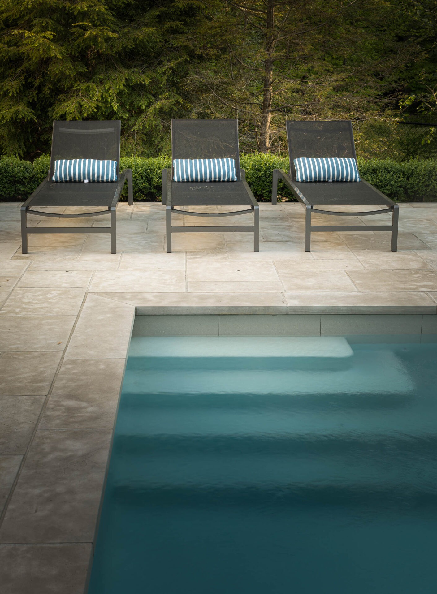 Three lounge chairs with striped pillows by a poolside, surrounded by lush greenery and stone tiles, creating a serene outdoor setting.