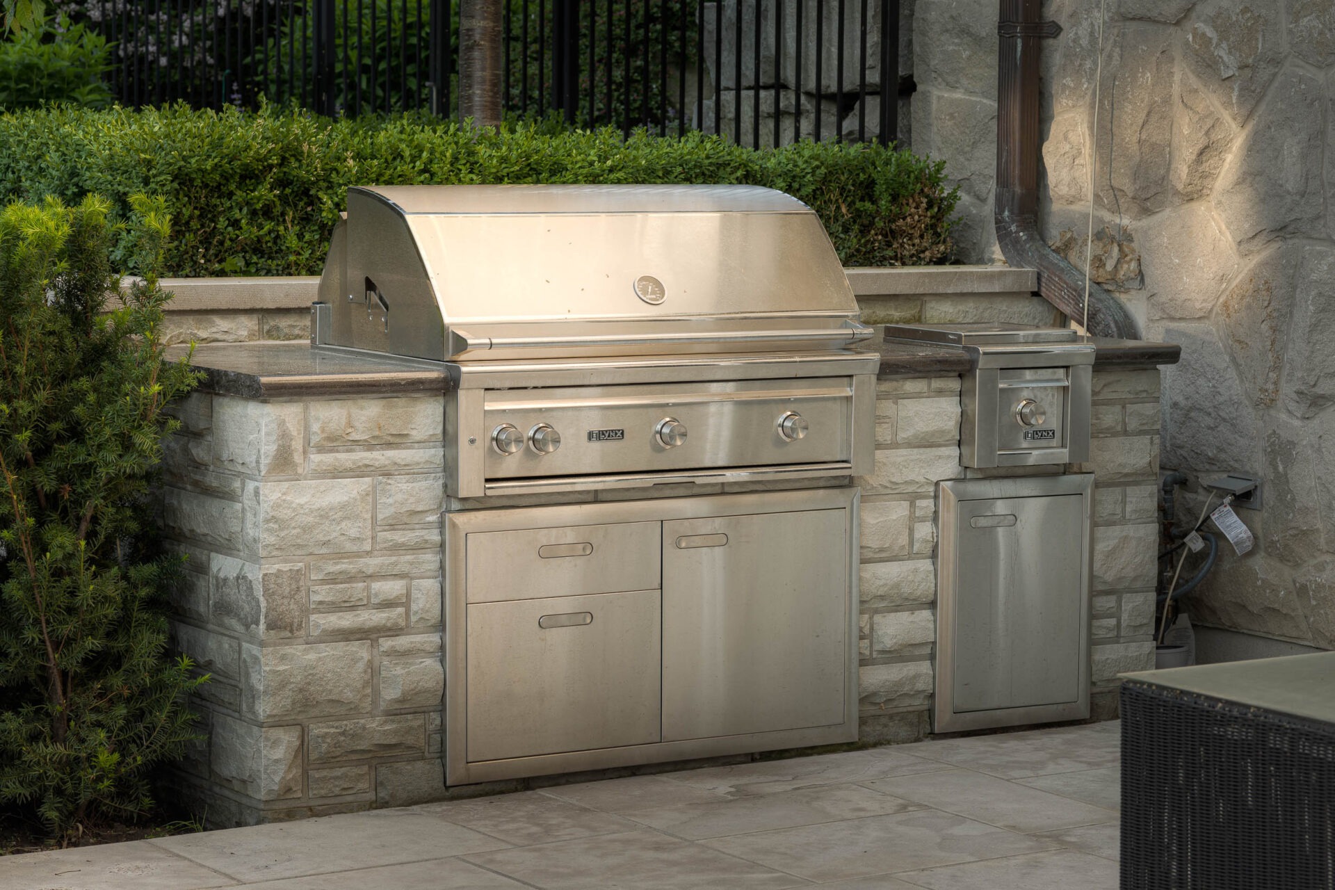 A stainless steel outdoor grill setup is surrounded by stone and greenery in a patio area, with a metal fence in the background.