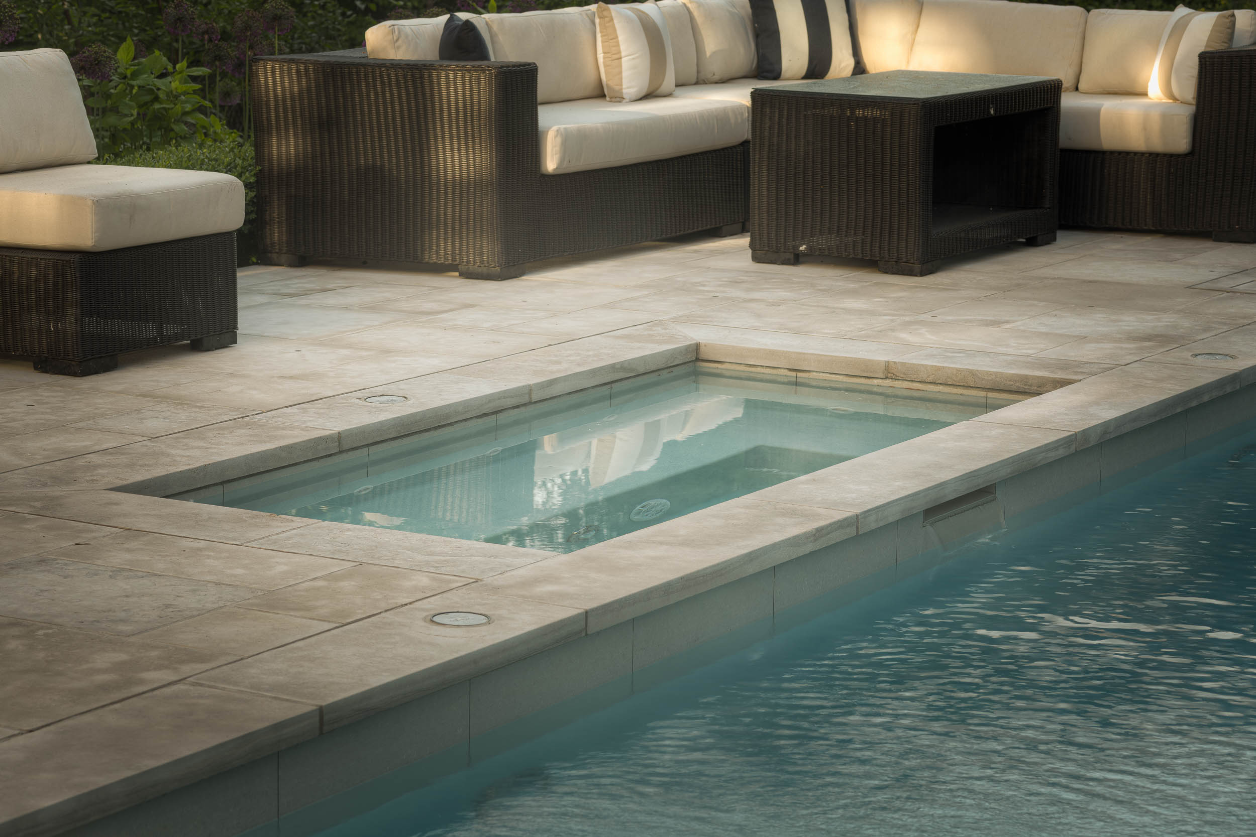 Cozy outdoor patio with wicker furniture and cushions, adjacent to a small, rectangular, turquoise swimming pool. Lush greenery surrounds the area.