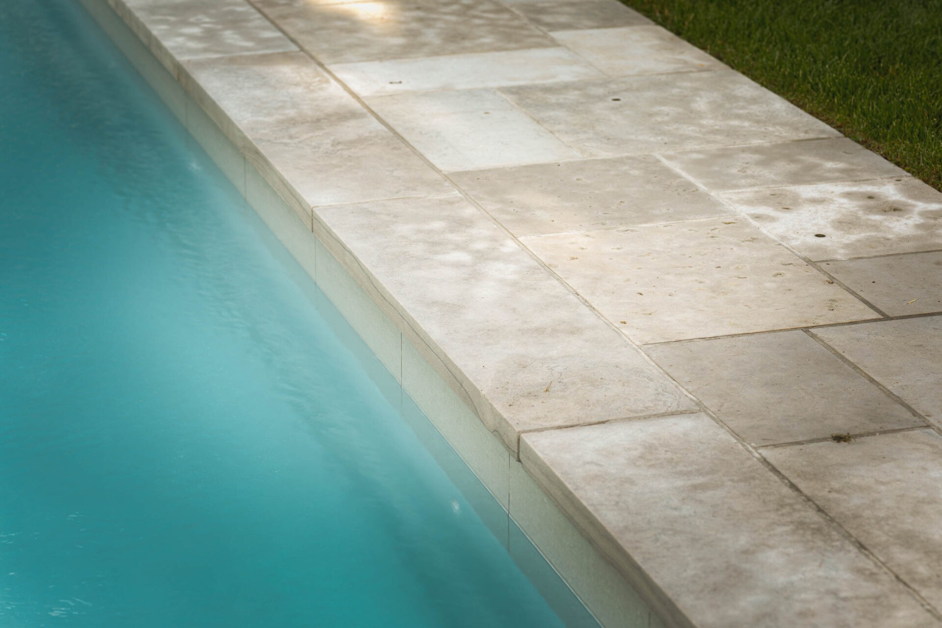 A tranquil pool edge features clean, light stone tiles bordering clear blue water, with adjacent green grass. Peaceful outdoor setting.