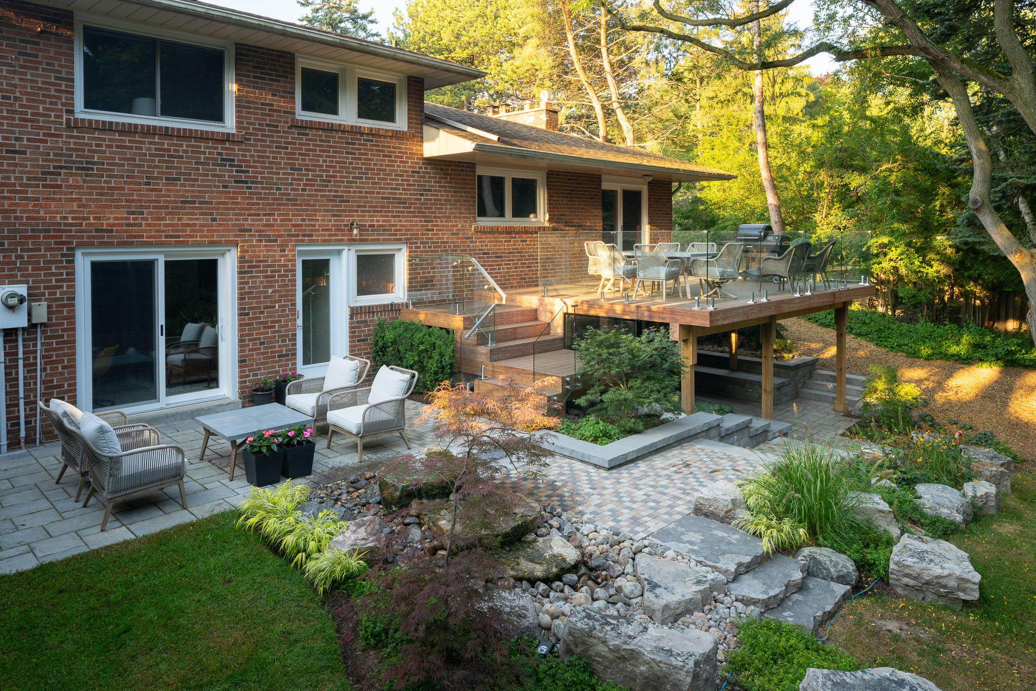 Brick house features patio with chairs, elevated deck, glass railing, lush landscaping, stone steps, and trees. Outdoor area designed for relaxation.