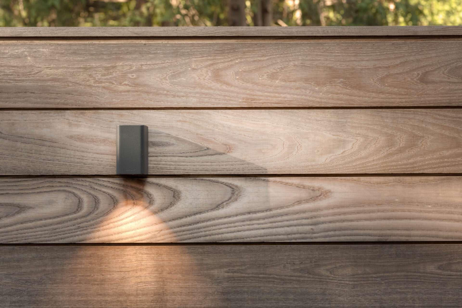 Wooden wall with a modern black light fixture casting a soft glow, surrounded by greenery in the background.