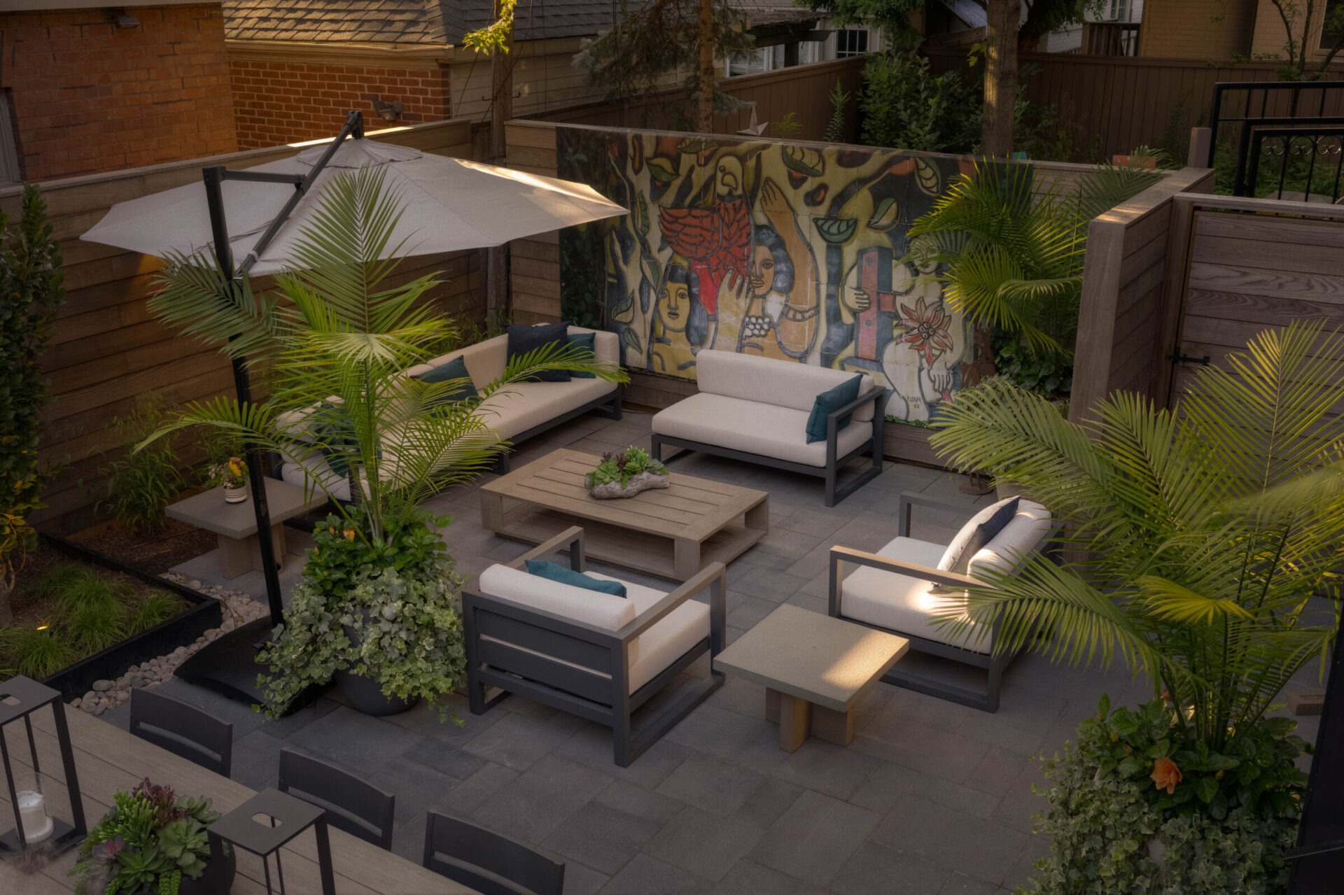 Cozy outdoor patio with modern gray furniture, potted plants, and a colorful mural on the wooden fence under a large umbrella.