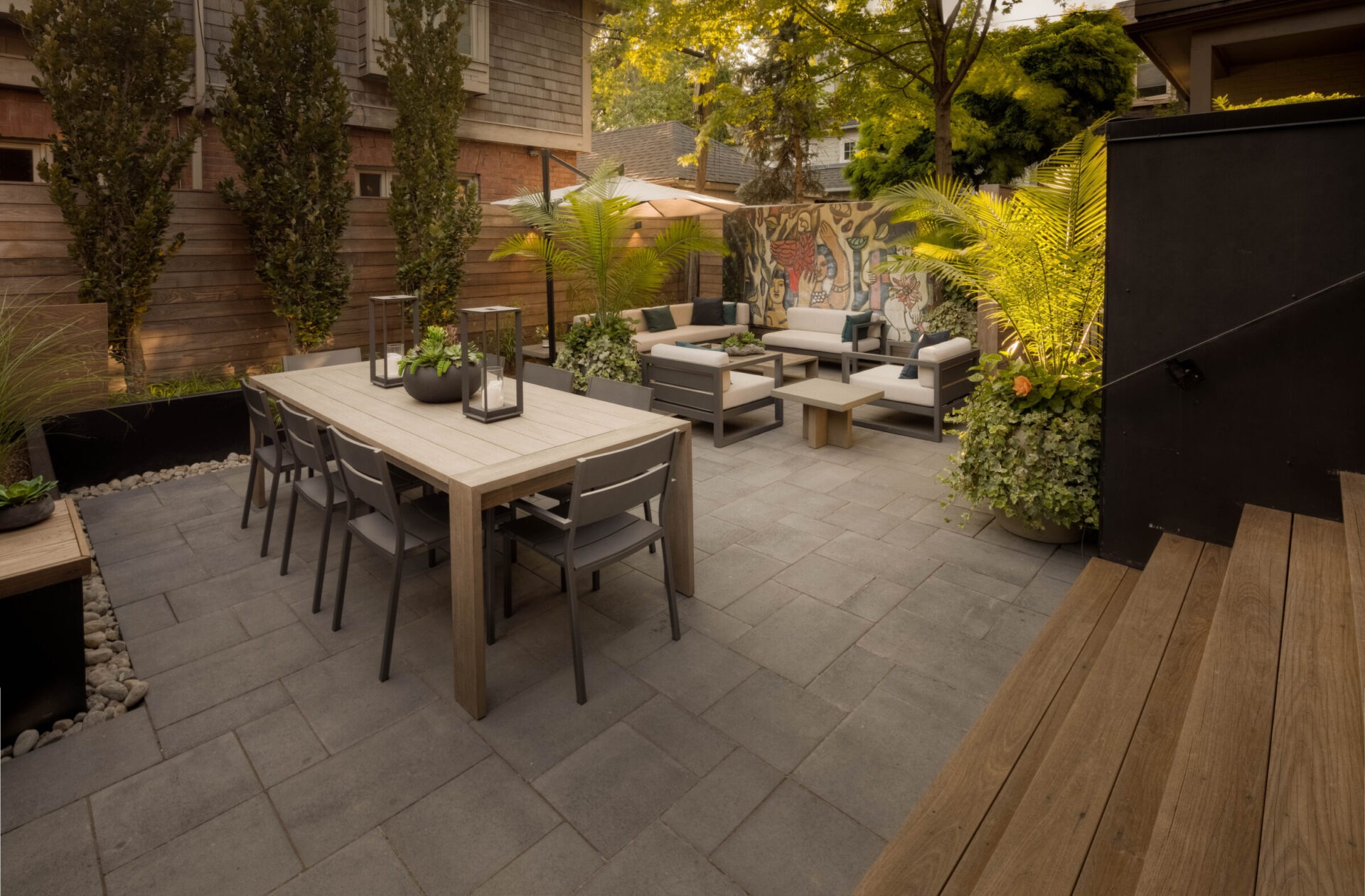 Modern patio with outdoor seating, dining table, and lush plants. Wooden fencing and vibrant mural enhance the cozy, stylish atmosphere, ideal for gatherings.