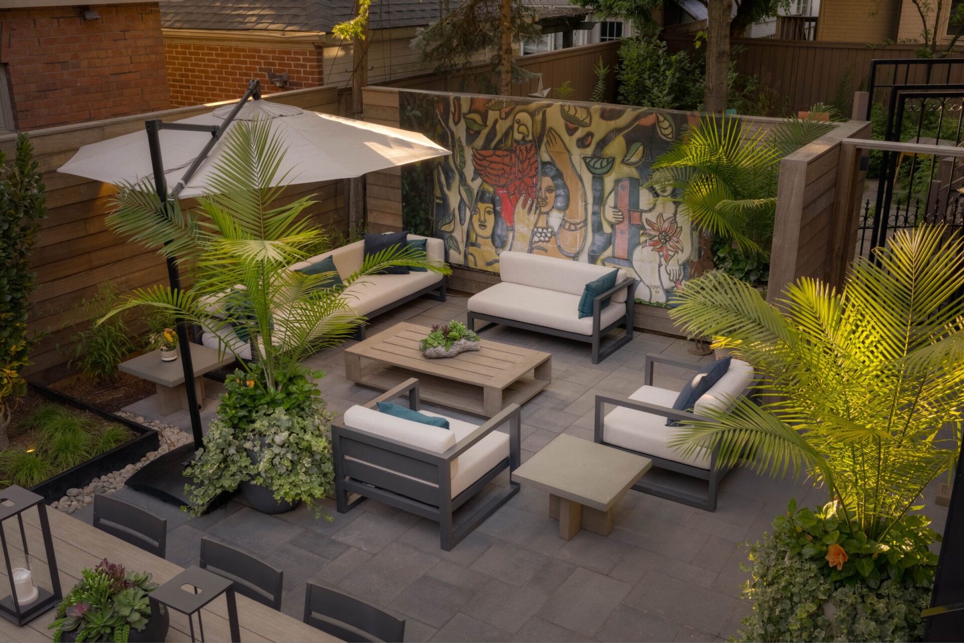 A cozy patio garden with modern furniture, potted plants, large umbrella, and artistic mural, surrounded by wooden fencing and urban buildings.