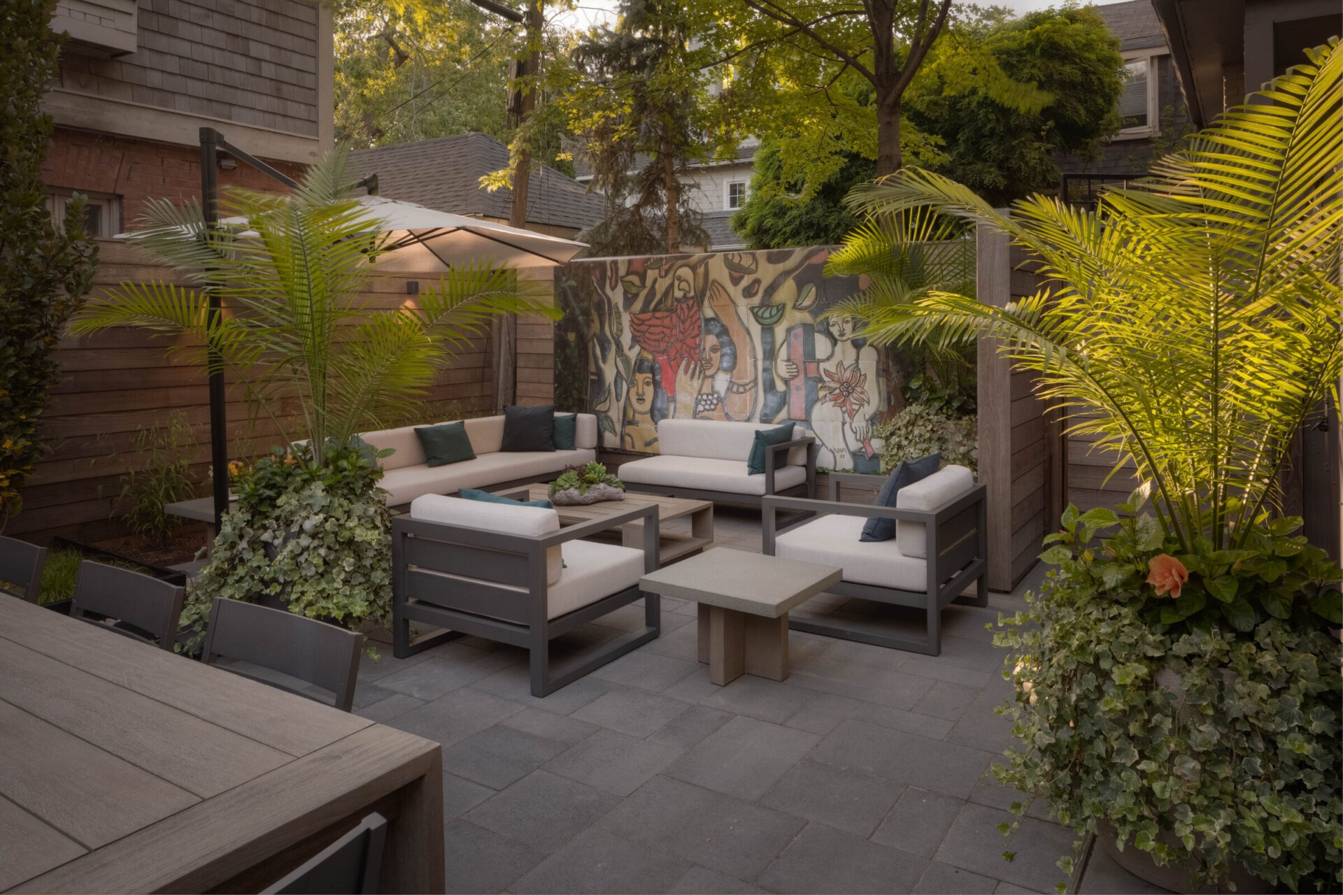 Cozy outdoor patio with modern furniture, vibrant mural, lush greenery, and umbrella, creating a serene and inviting atmosphere for relaxation and gatherings.