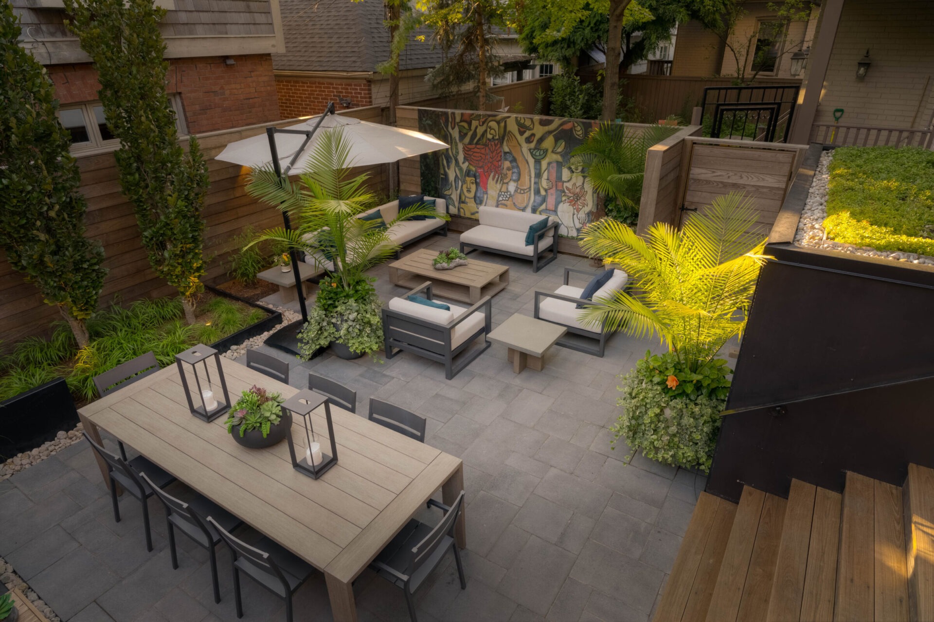 Contemporary patio with seating and dining areas, surrounded by lush plants. Wall features colorful artwork, creating a vibrant, inviting outdoor space.