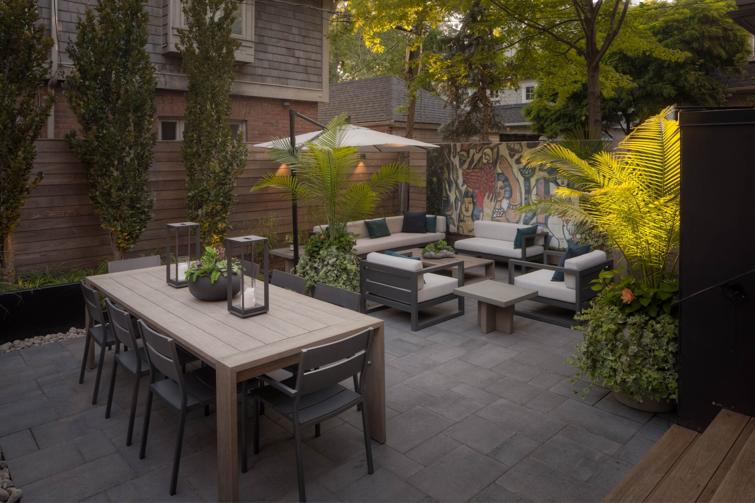 Contemporary patio with modern furniture, green plants, and mural art. Wooden table, umbrellas, and soft lighting create a cozy outdoor setting.