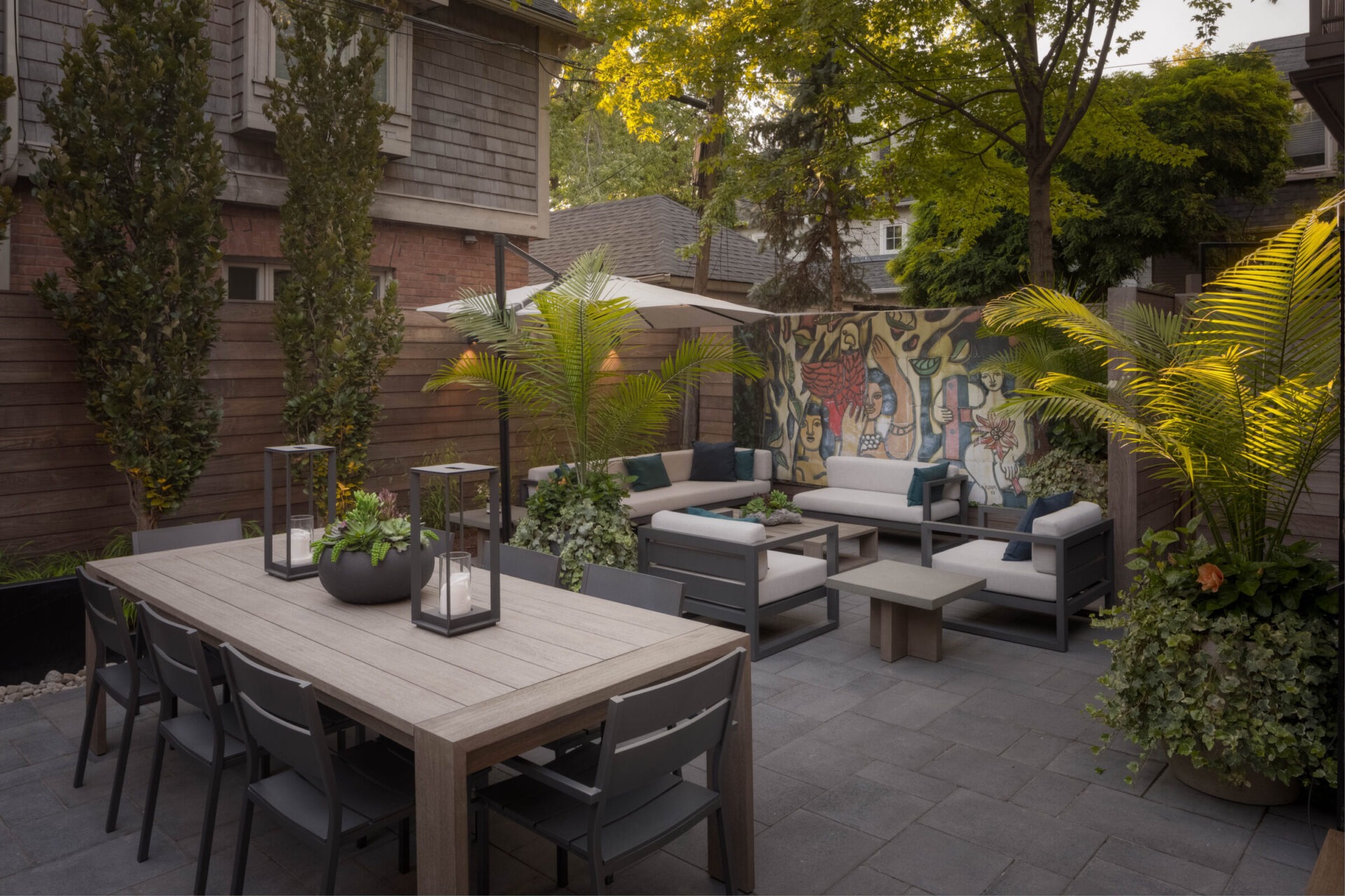 Charming outdoor patio with modern dining set, lush plants, comfortable seating, and vibrant mural. Cozy atmosphere surrounded by wooden fencing and greenery.