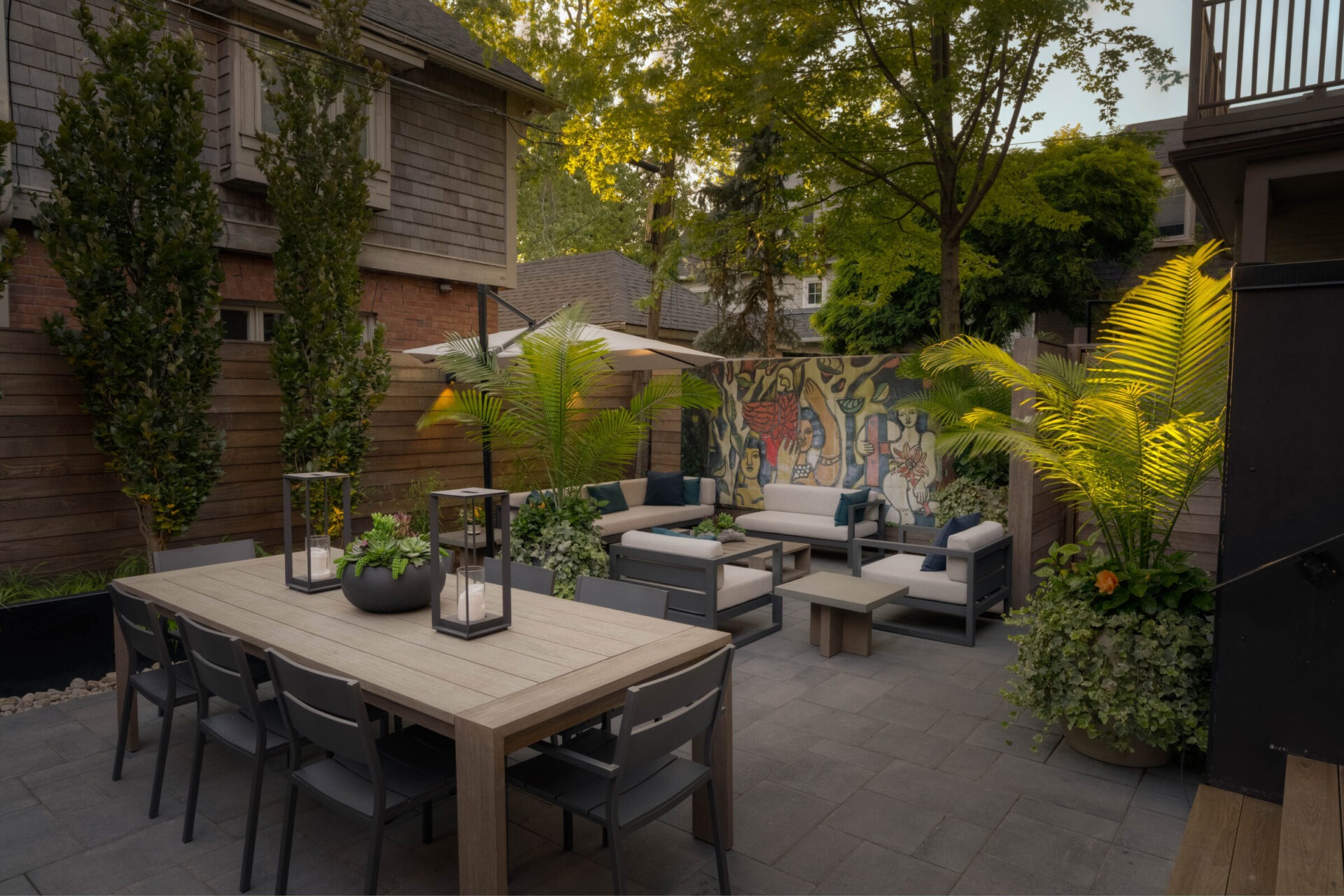 Modern backyard patio with sectional sofas, dining table, and potted plants. Colorful mural on wooden fence adds artistic flair to the setting.