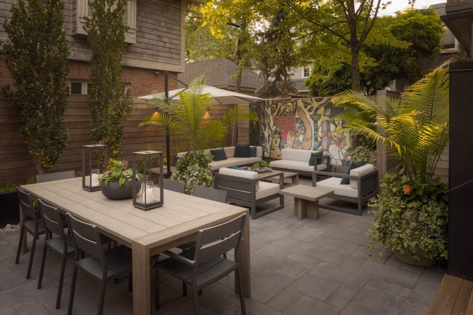 Cozy backyard patio with modern furniture, lush greenery, large dining table, and colorful mural on wooden fence, surrounded by trees and brick building.