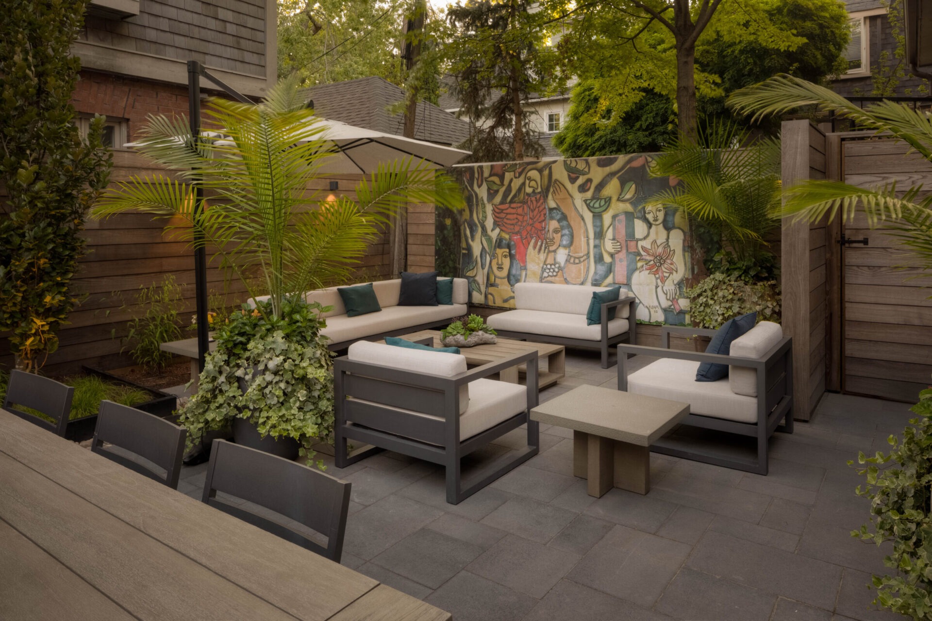 A cozy outdoor seating area with modern furniture, vibrant mural, lush plants, in a private garden setting surrounded by wooden fencing.