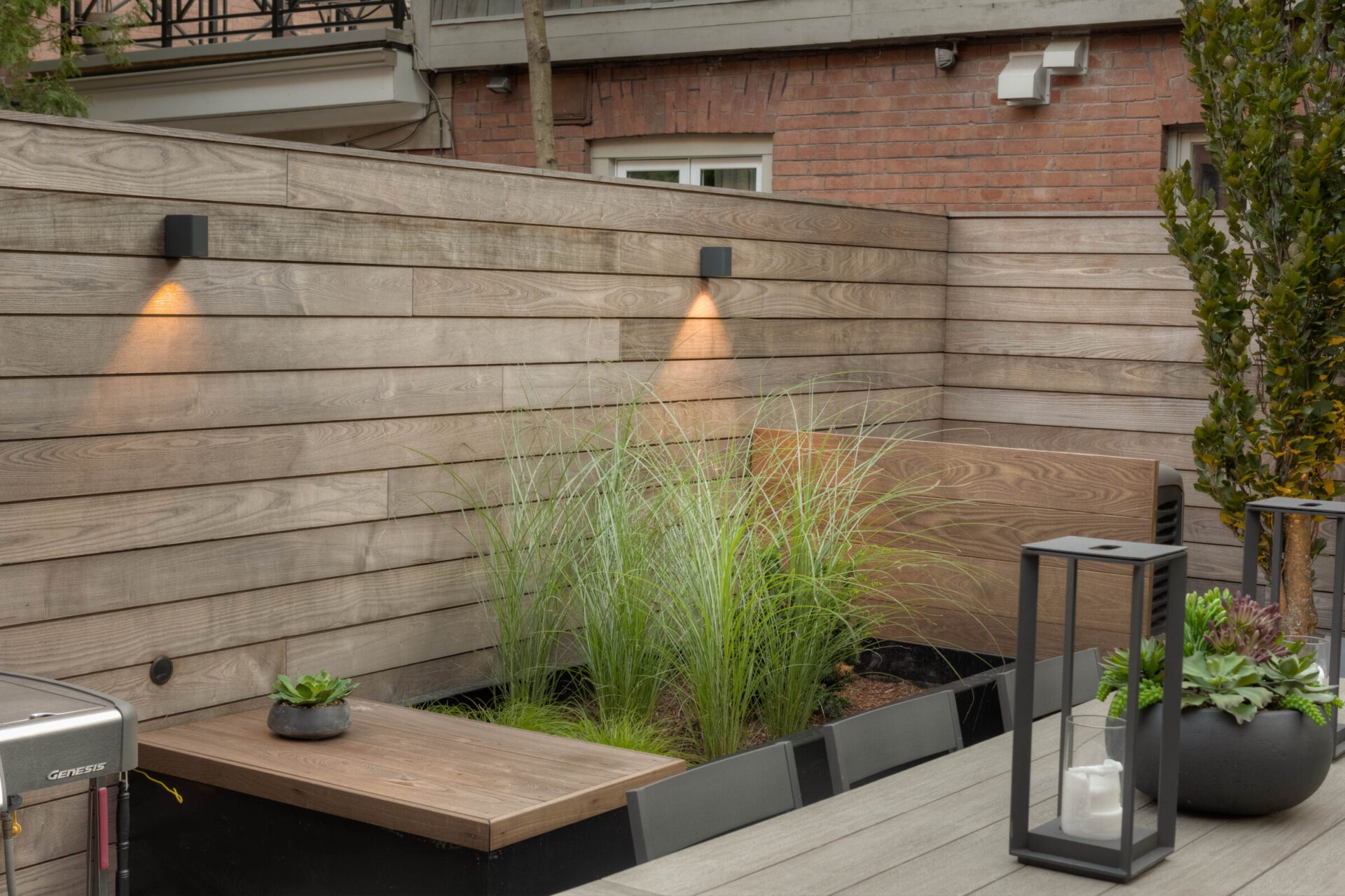 Modern outdoor patio with wooden paneling, decorative lighting, lush greenery, and minimalist planters. A small seating area complements the tranquil setting.