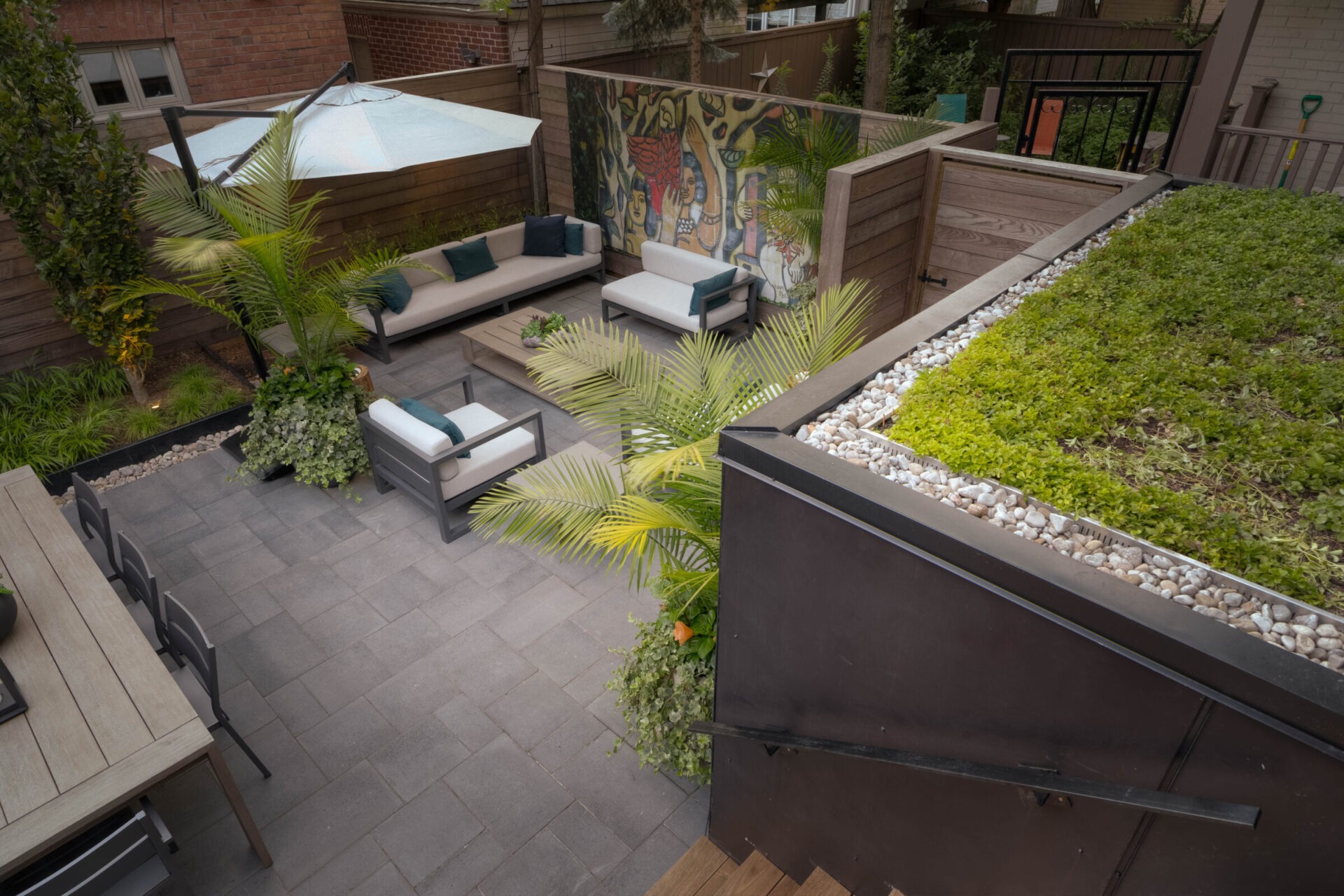 Modern patio with seating, plants, and a mural. Elevated view reveals greenery on a rooftop. Peaceful outdoor space for relaxation and gatherings.