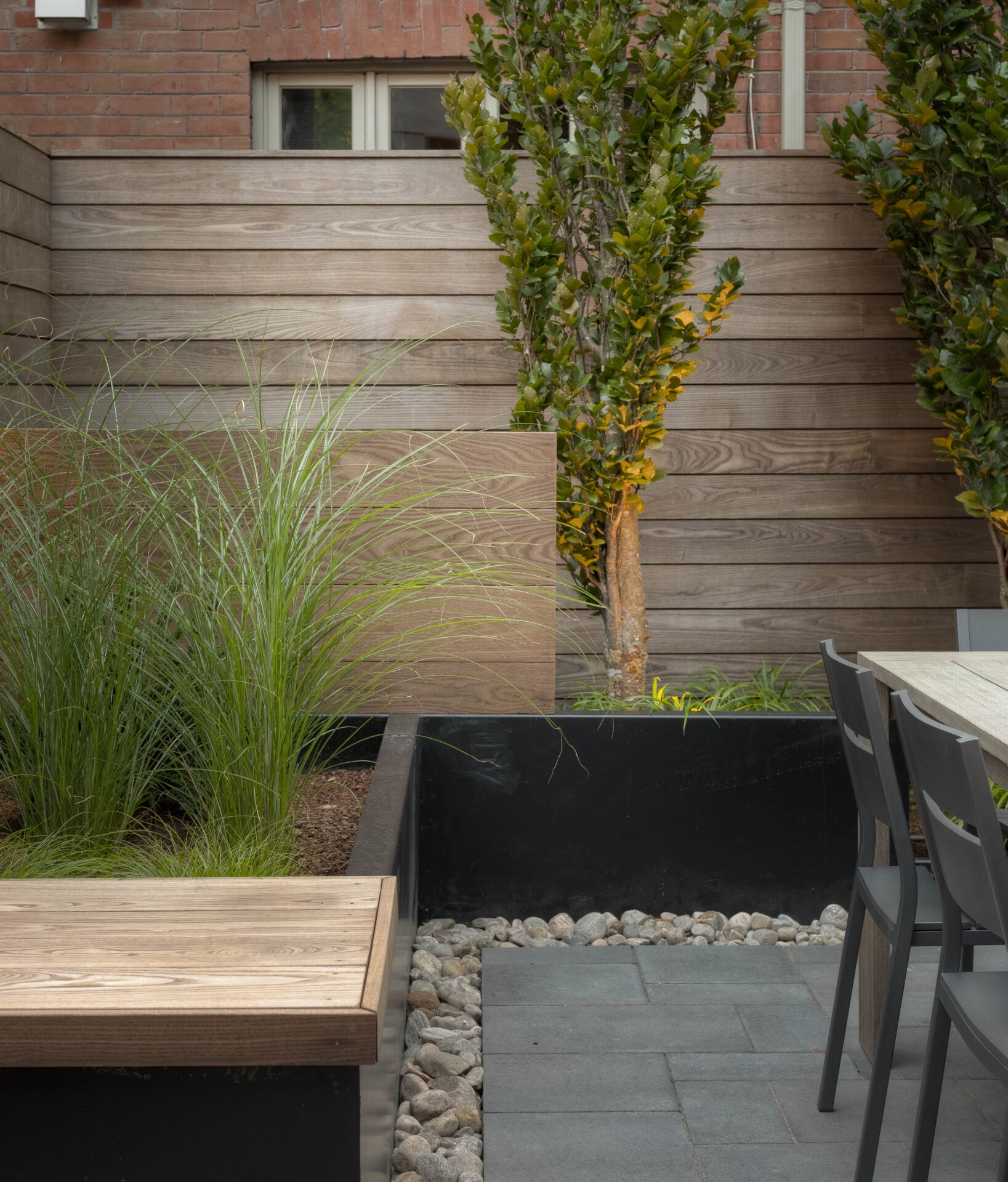 A modern garden features wooden fencing, tall grasses, trees, a bench, and dining chairs, creating a peaceful outdoor seating area.