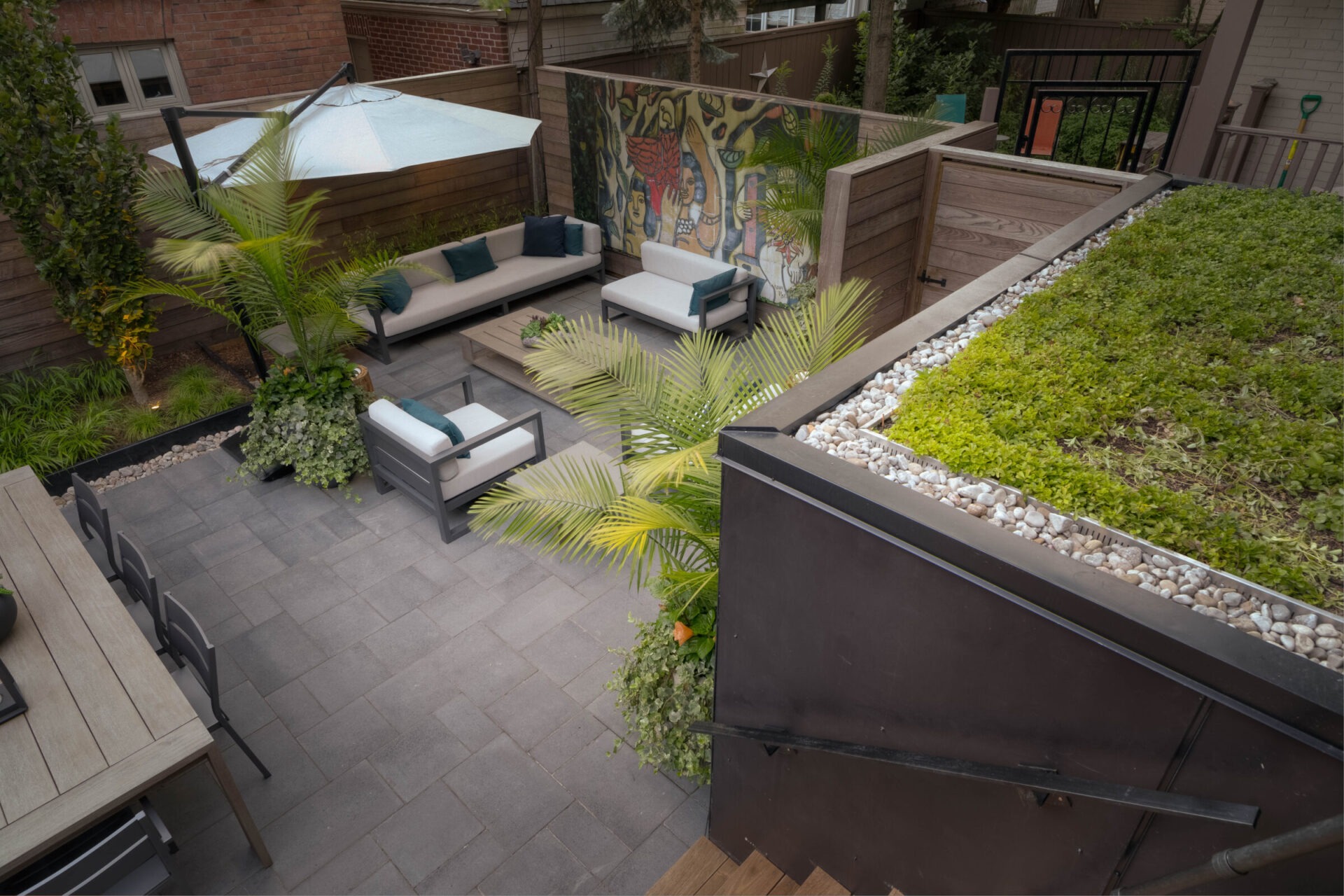 Modern backyard patio with seating, umbrella, and vibrant mural. Green rooftop garden and potted plants add lush accents to the space.