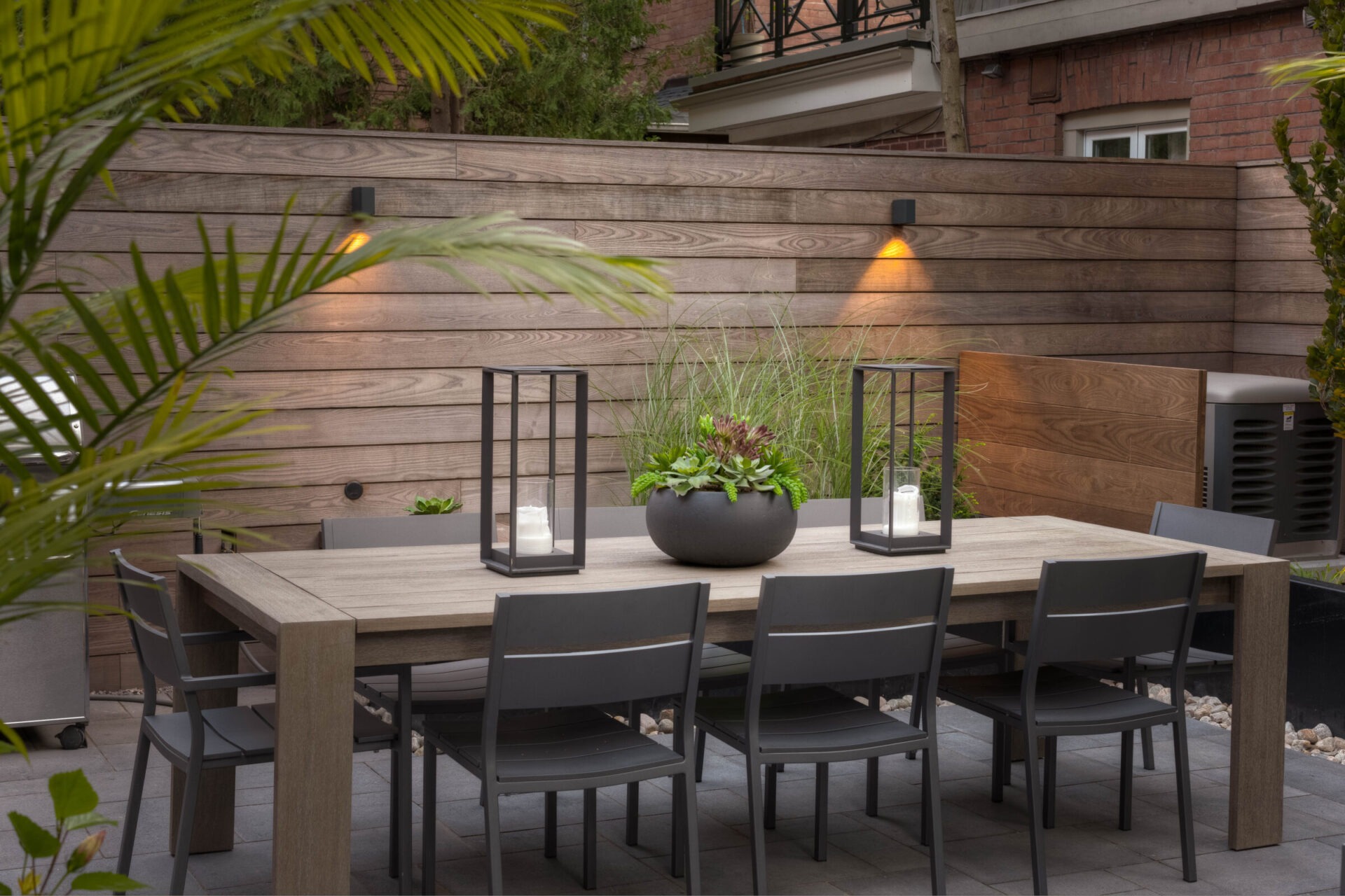 Modern outdoor patio with a wooden dining table, black chairs, decorative lanterns, plants, and soft lighting against a wood-paneled fence.