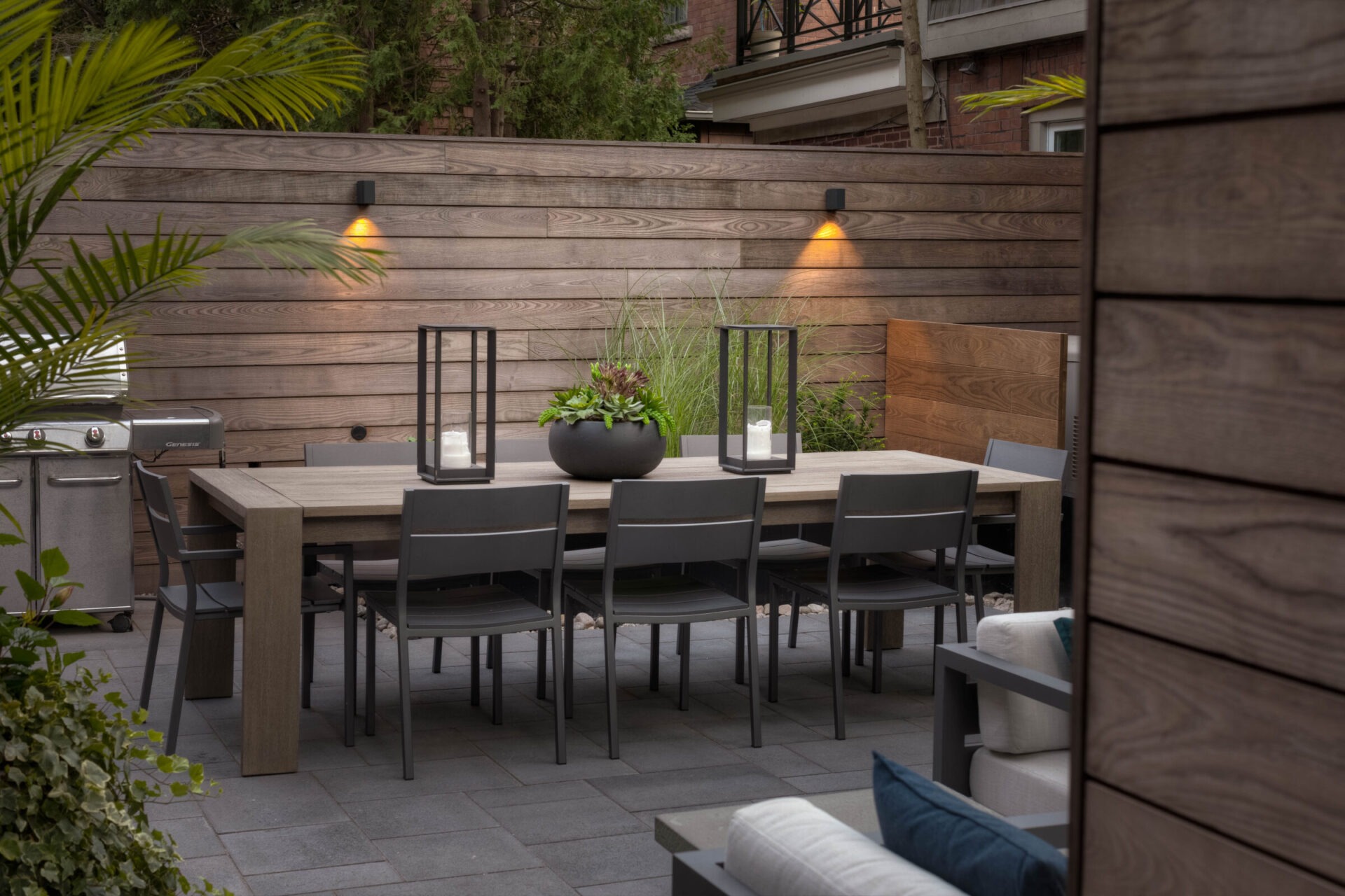 Modern patio with wooden walls, a large dining table, eight chairs, decorative plants, lanterns, ambient lighting, and a stainless steel grill.