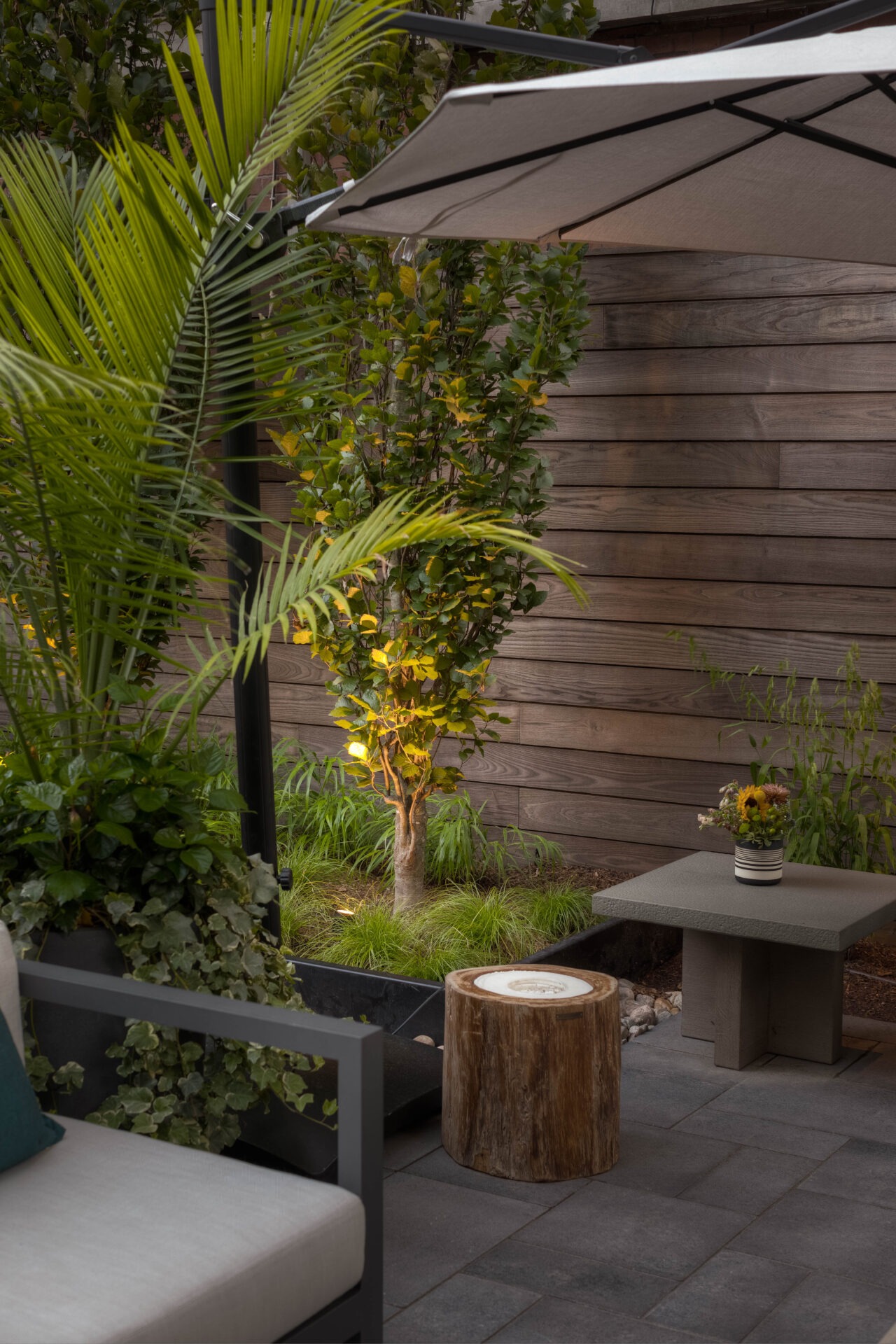 A cozy patio with potted plants, a small tree, wooden stool, and outdoor furniture under a shade, surrounded by a wooden fence.