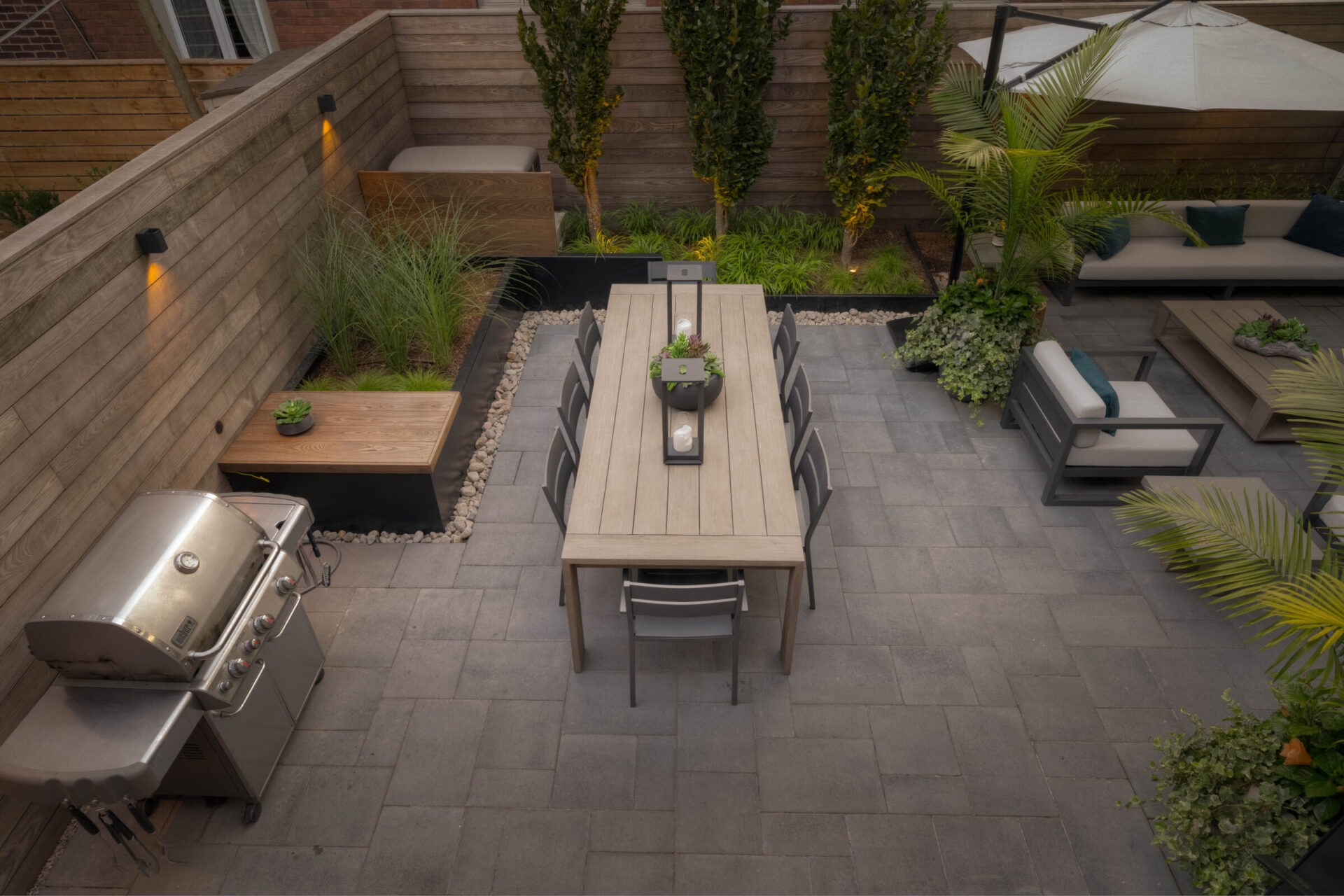 Modern patio with dining table, chairs, BBQ grill, plants, and cozy seating. Enclosed by wooden fencing, creating a serene outdoor space.