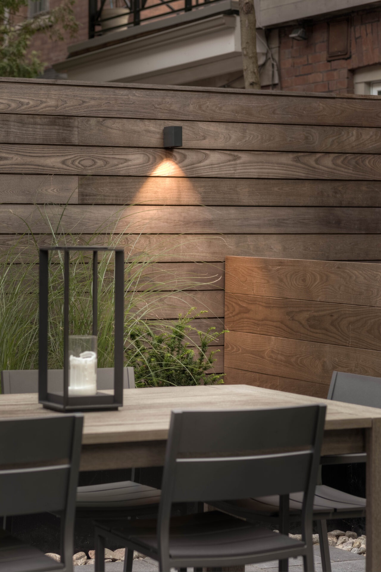 Modern outdoor patio with wooden fence, table, and chairs. Soft lighting highlights green plants and a candle lantern centerpiece on the table.