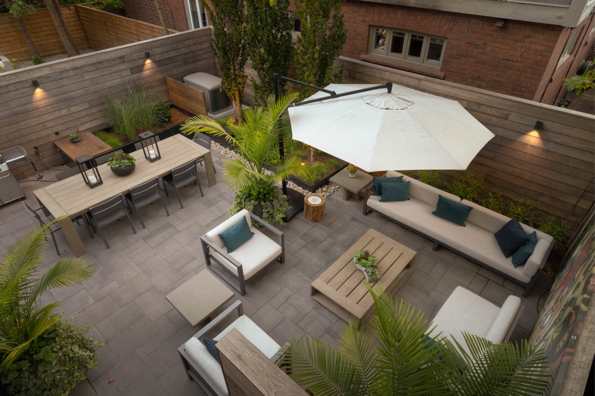 Outdoor patio with seating area, dining table, umbrella, and potted plants. Modern design with wooden elements, surrounded by tall brick and wooden walls.