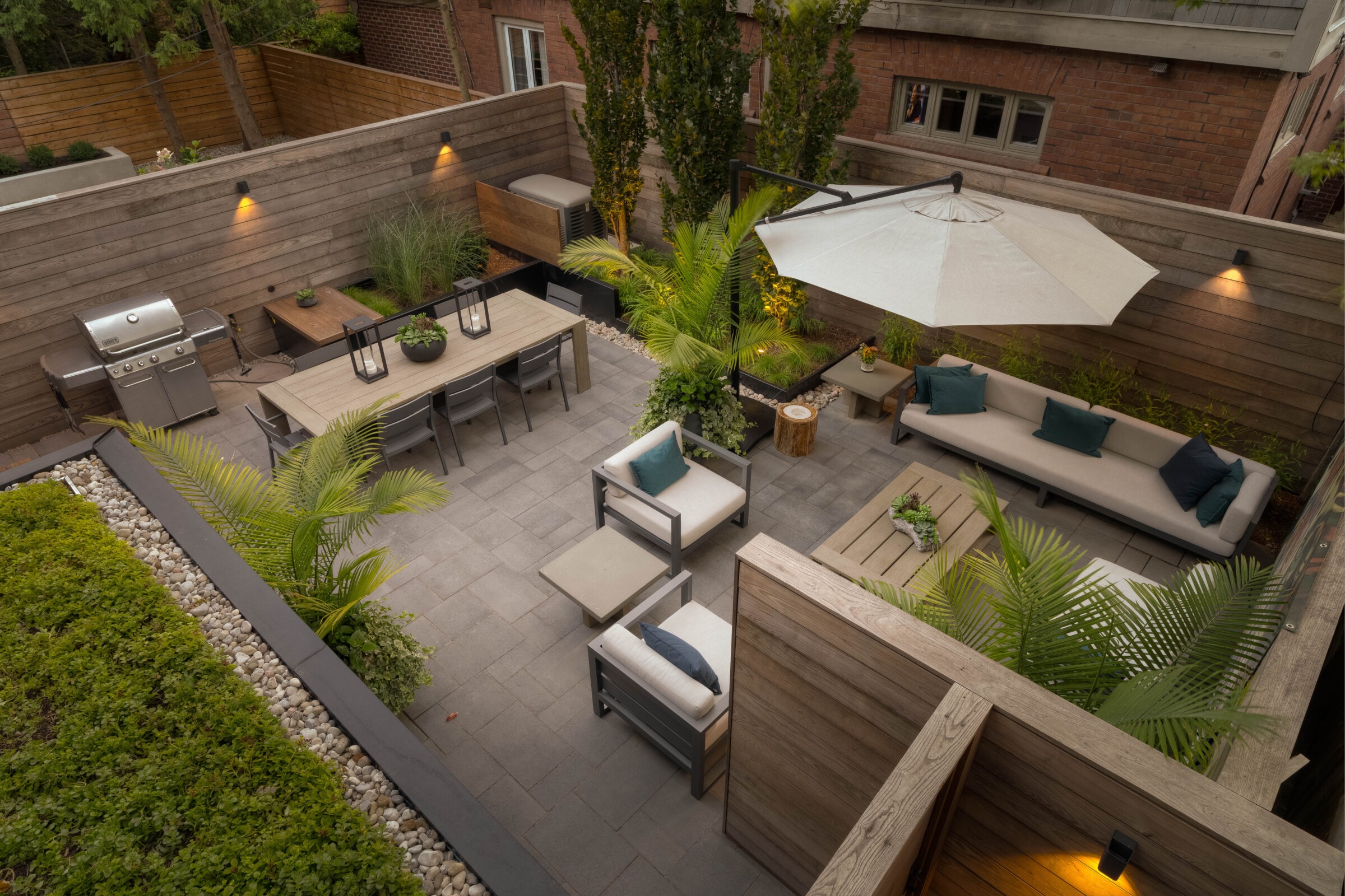 Modern patio with gray stone tiles, wooden furniture, potted plants, large umbrella, grill, and privacy walls surrounded by lush greenery and trees.
