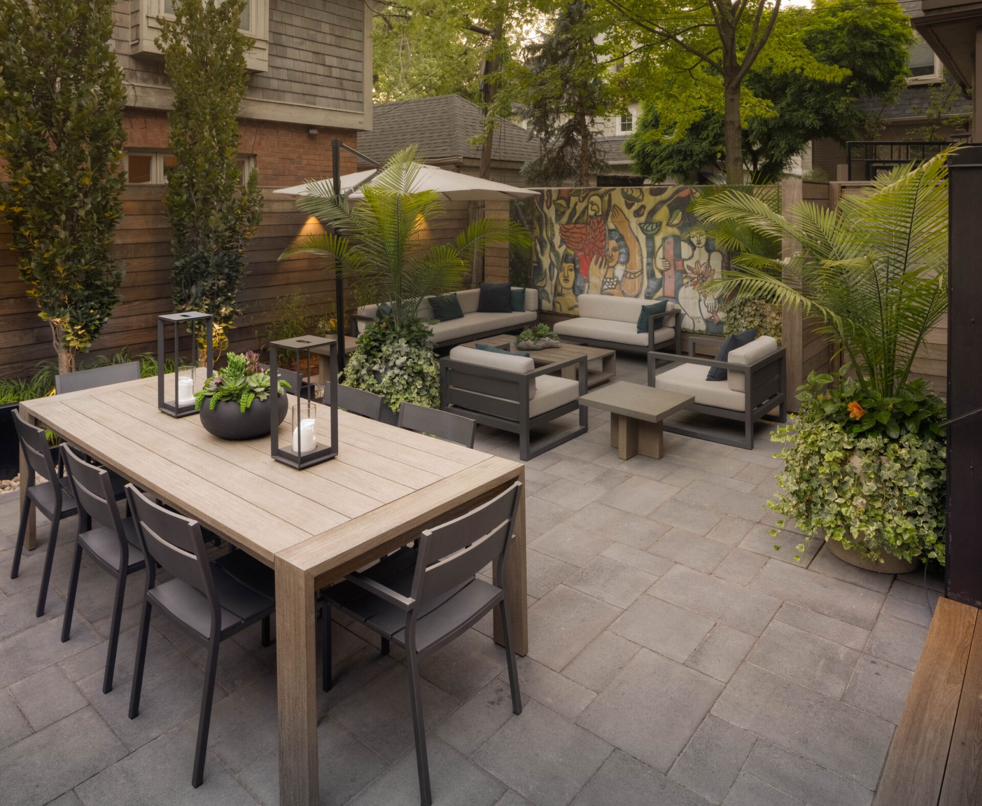 Modern backyard patio with wooden dining table, chairs, and comfortable seating area. Artistic mural on the back wall, surrounded by lush greenery.