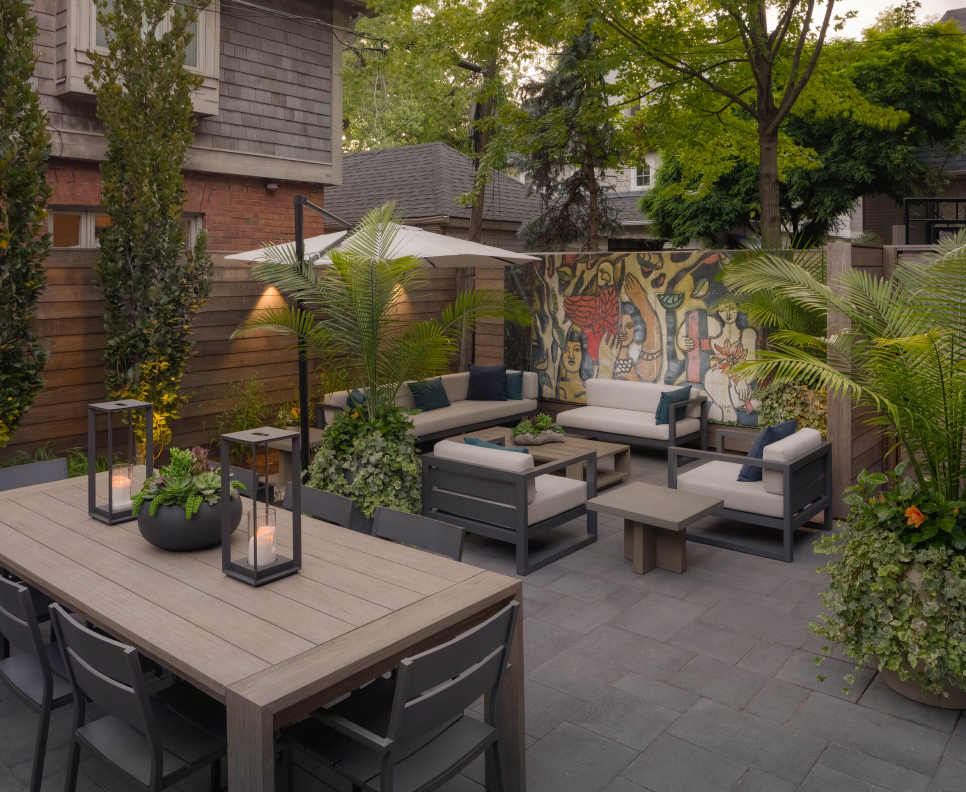 A cozy, modern outdoor patio with seating, tropical plants, a large mural, and a wooden dining table, surrounded by trees and fencing.