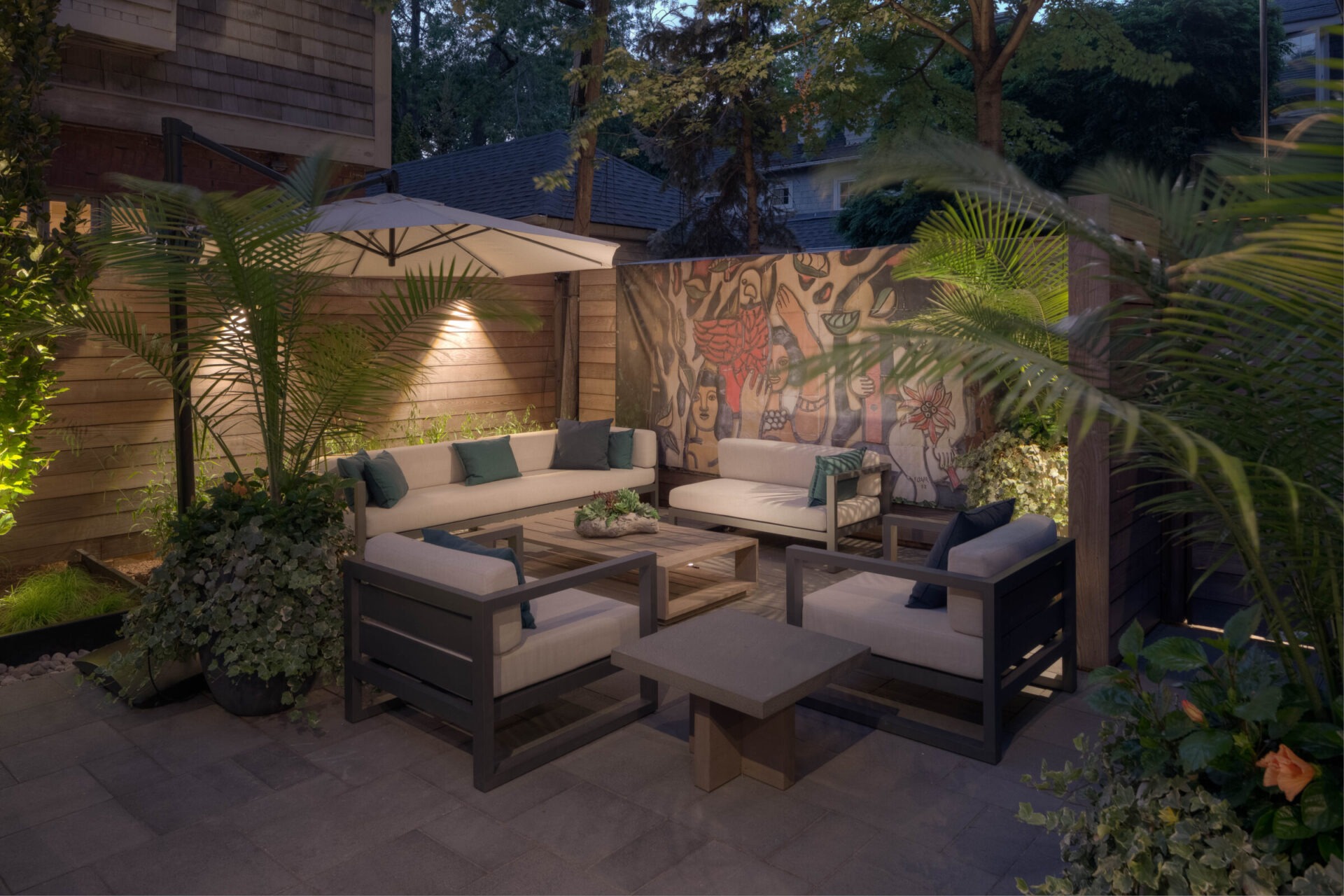 Cozy outdoor patio with contemporary furniture, an umbrella, and artistic mural surrounded by lush plants and wooden fencing, creating a serene atmosphere.