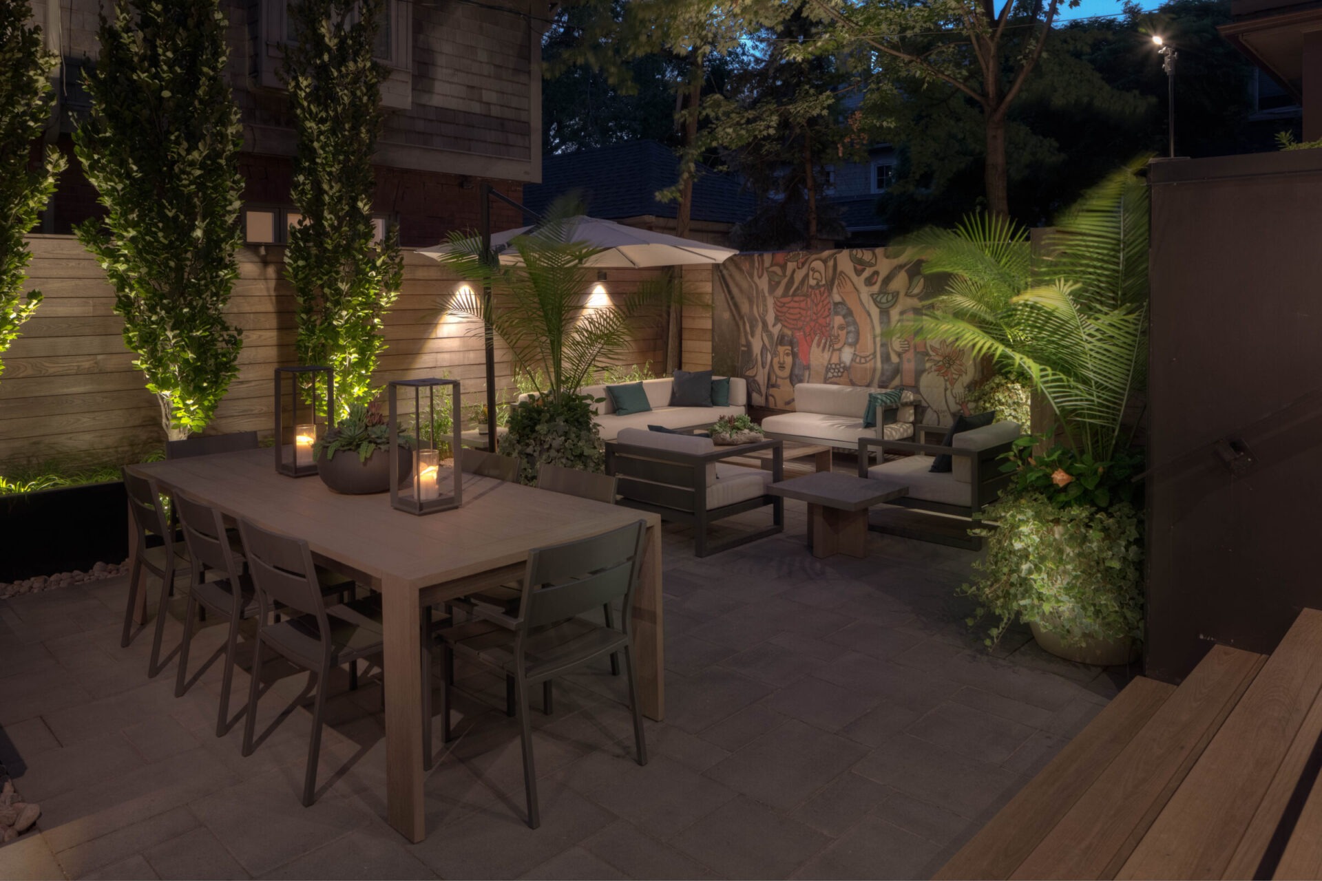 Outdoor patio with a wooden table, chairs, sofas, potted plants, candles, and mural artwork on one wall, under evening lighting.