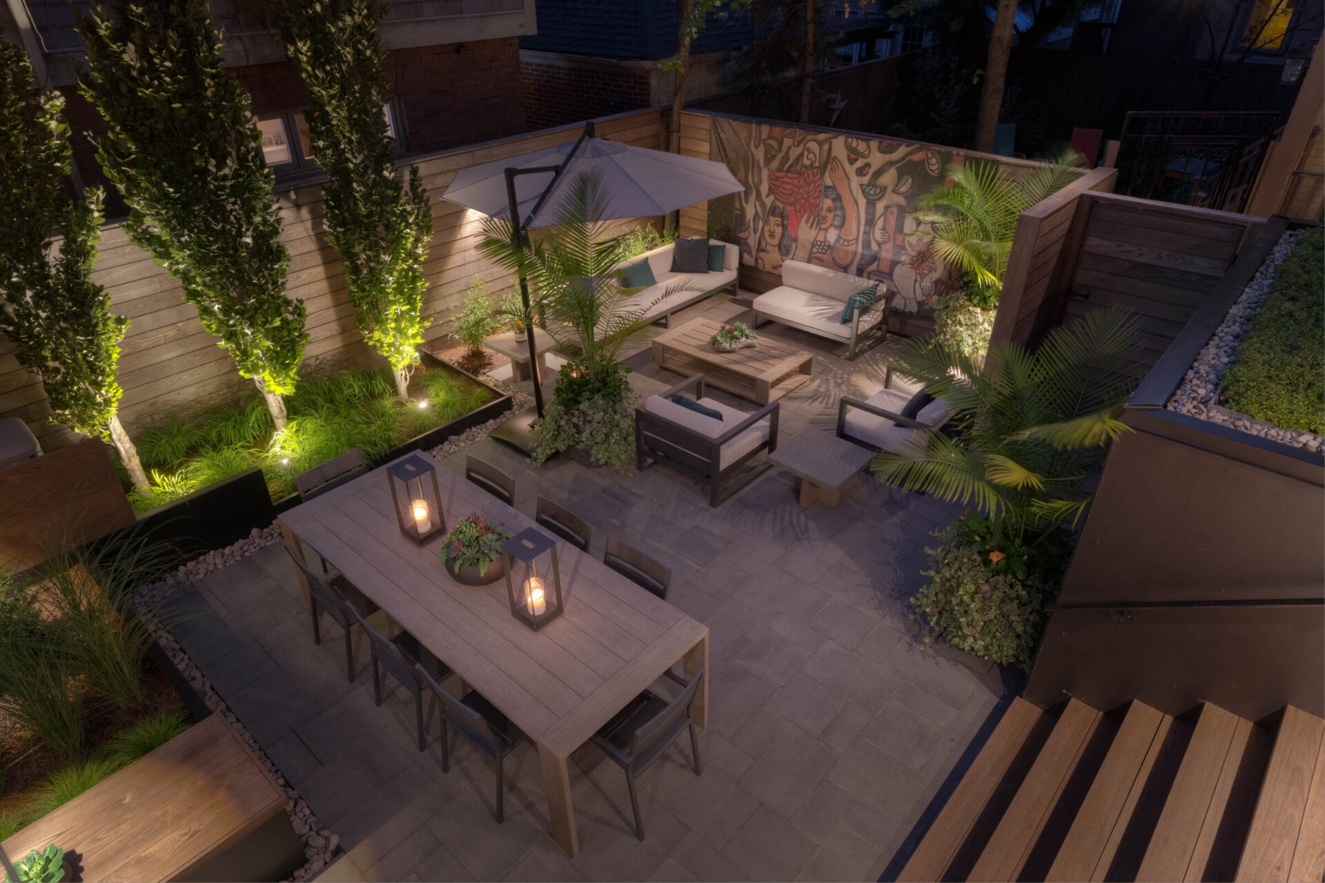 Modern outdoor patio with sofas, dining table, illuminated greenery, and artistic mural. Cozy ambiance with lanterns and umbrella under evening light.