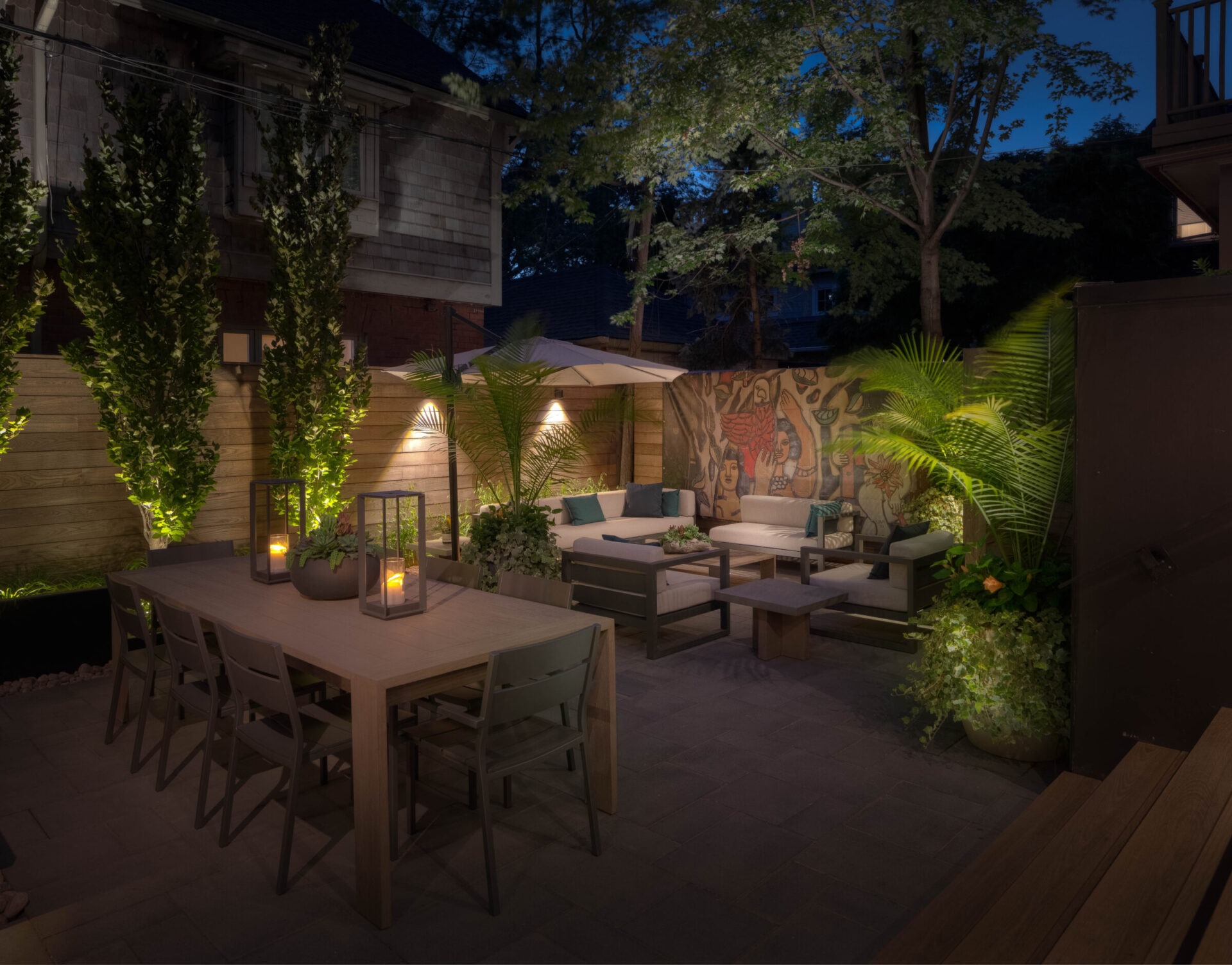 Cozy backyard with patio furniture, dining table, potted plants, and lanterns. Mural on wall enhances the intimate evening setting.