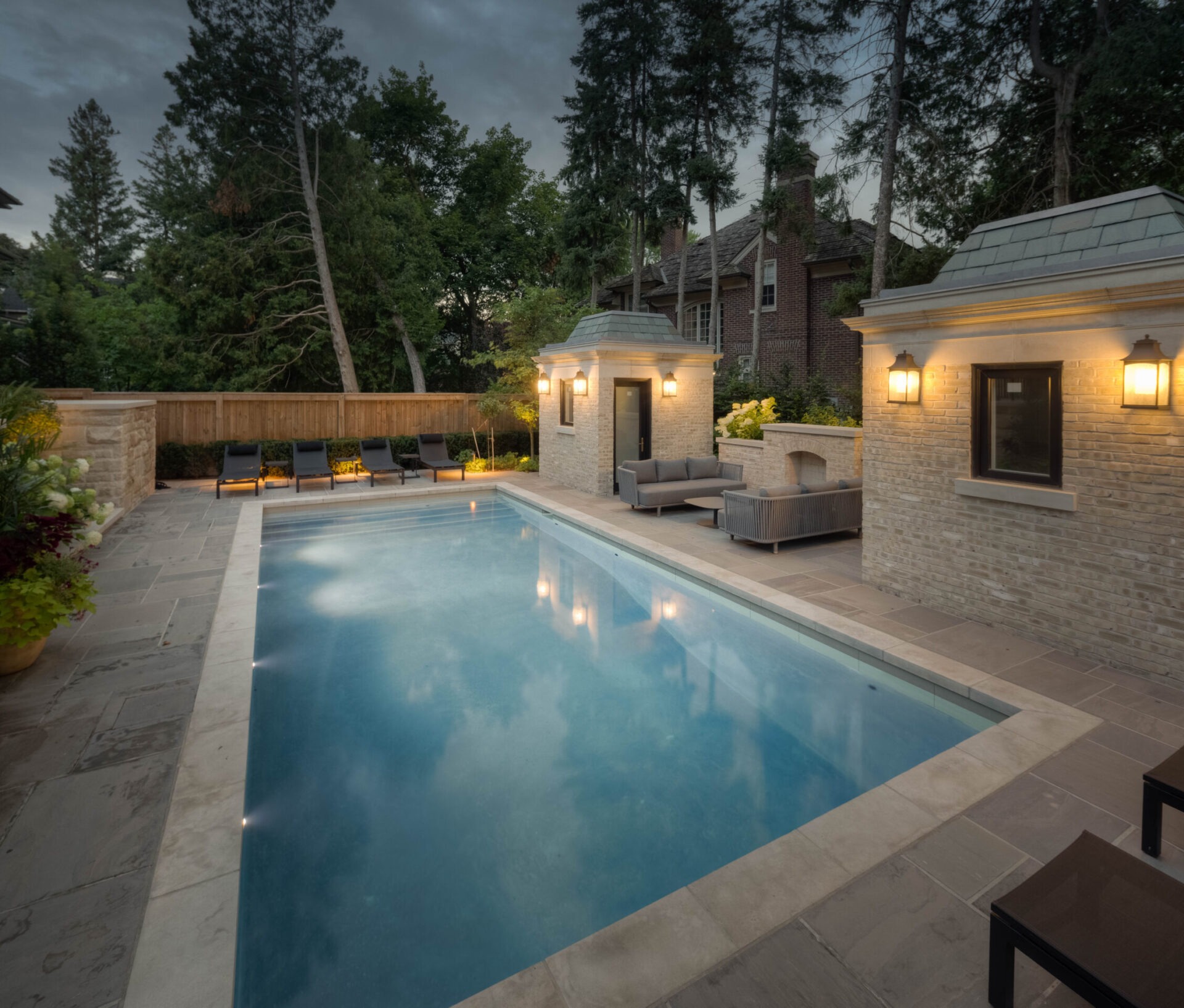 Elegant outdoor pool area surrounded by serene trees and comfortable seating, enhanced by warm lighting and a stylish fence.