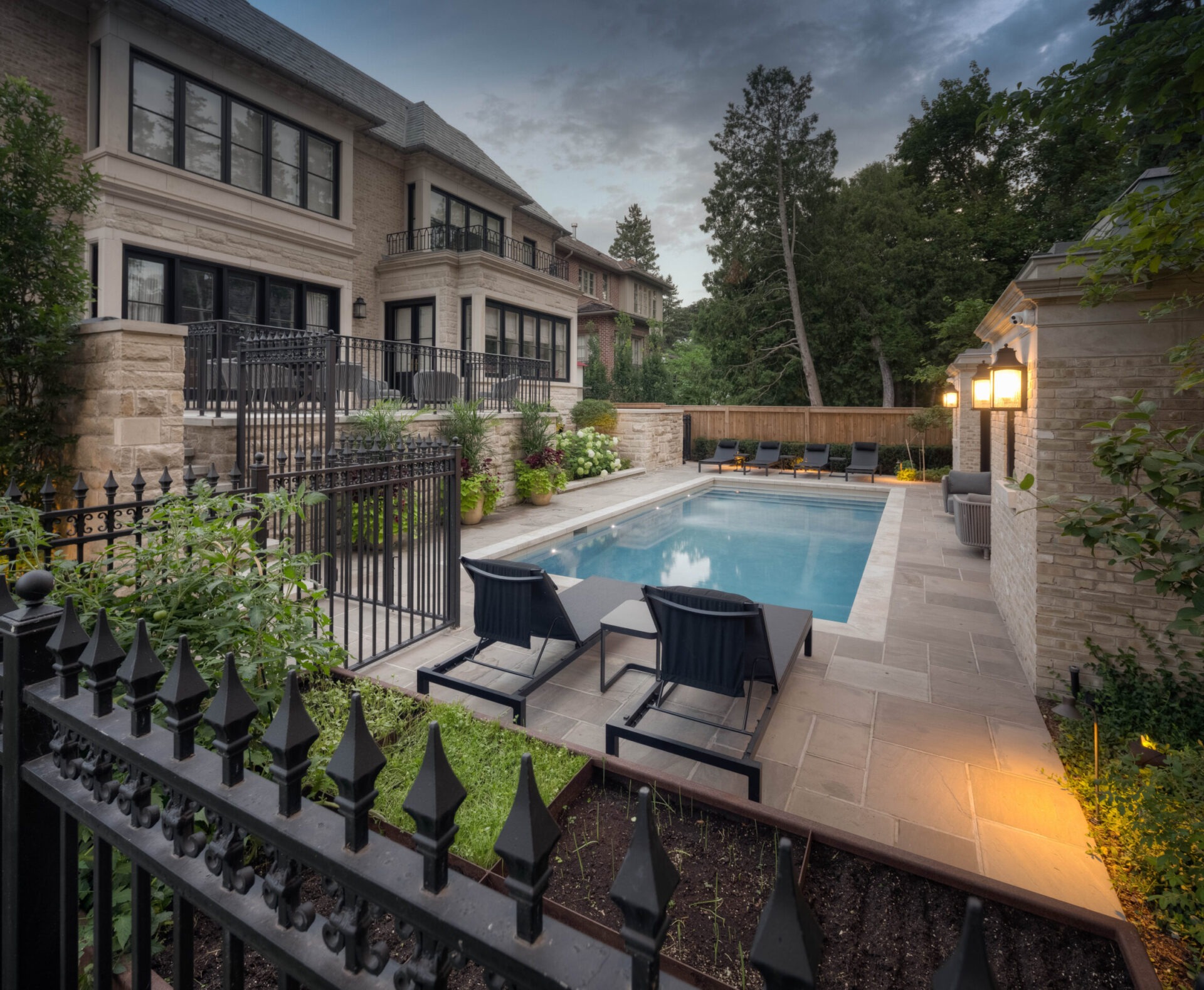 Elegant backyard with a rectangular pool, surrounded by modern patio furniture, lush greenery, and a large, sophisticated brick house at dusk.