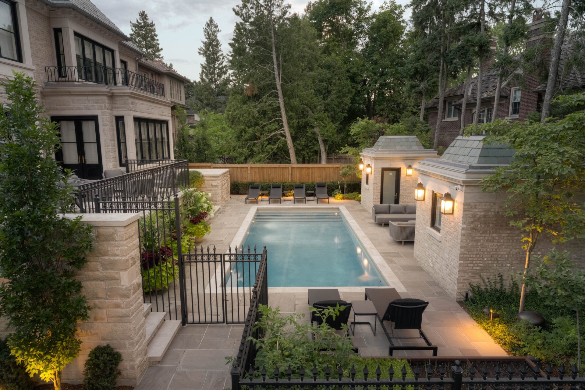 Luxurious backyard features a rectangular swimming pool, surrounded by elegant stone architecture, lush greenery, lounge chairs, and warm outdoor lighting.