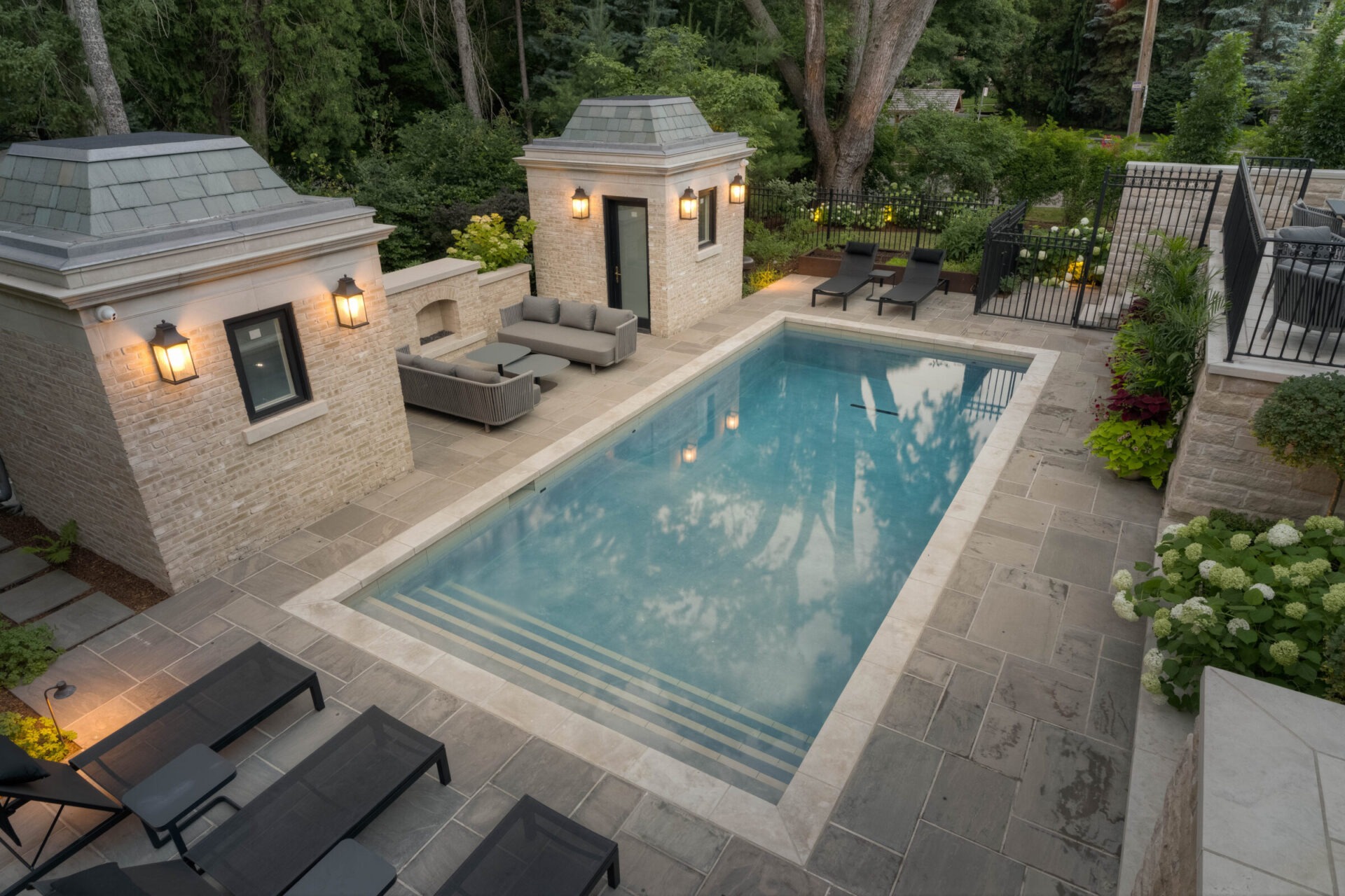 A serene backyard with a rectangular pool, surrounded by comfortable seating, lush greenery, and two brick buildings with warm exterior lighting.