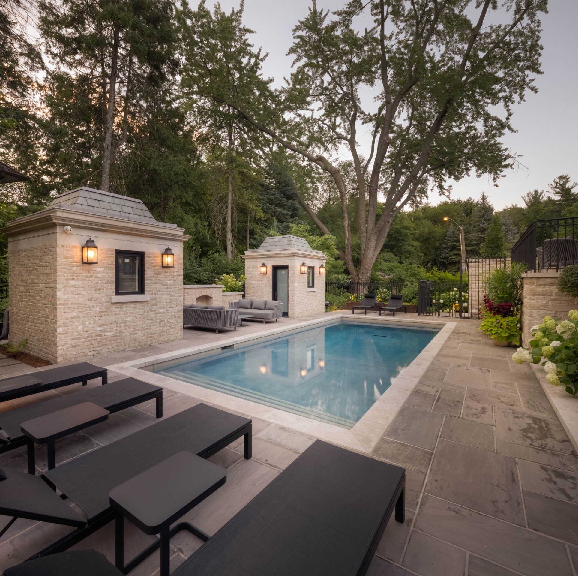 A serene backyard features a small pool, lounge chairs, and cozy seating areas, surrounded by lush greenery and two elegant brick structures.