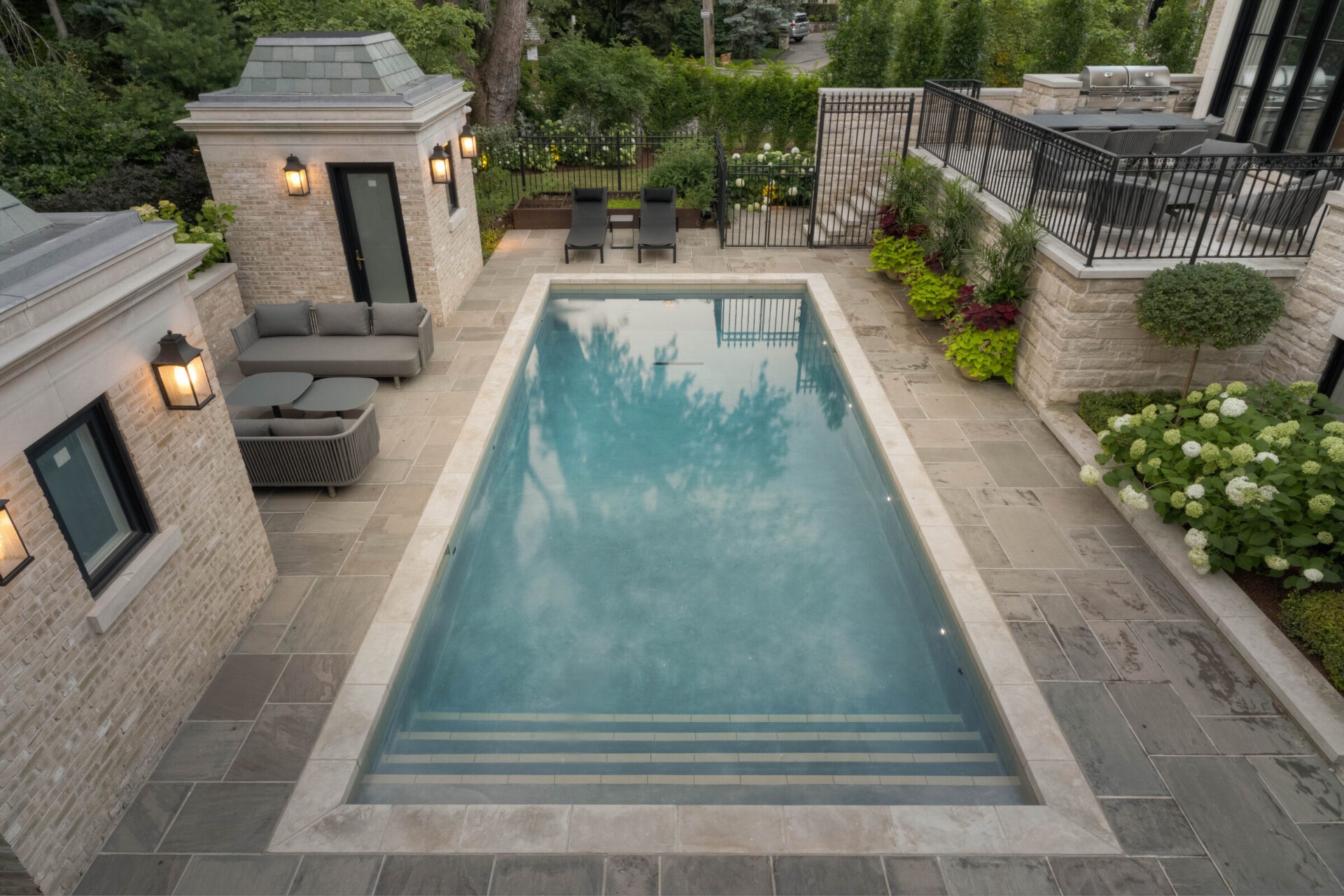 A modern rectangular pool surrounded by stone tile, patio furniture, lush greenery, and brick structures with lanterns in a landscaped garden.